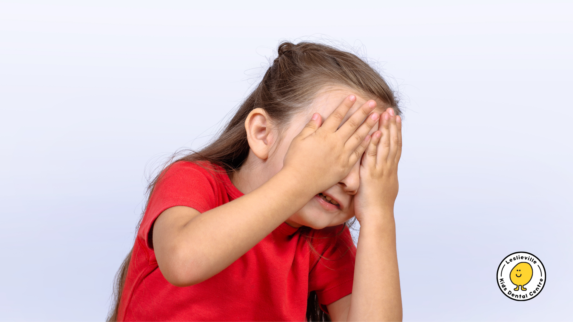 A person in a red shirt covering their face with their hands against a light background.