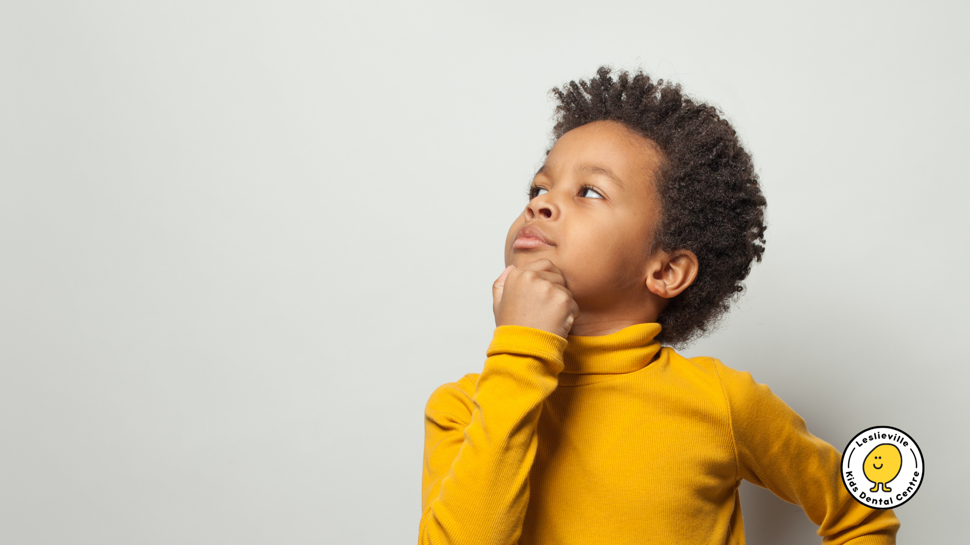 Boy in yellow turtleneck, hand on chin, looking upwards thoughtfully. Gray background.