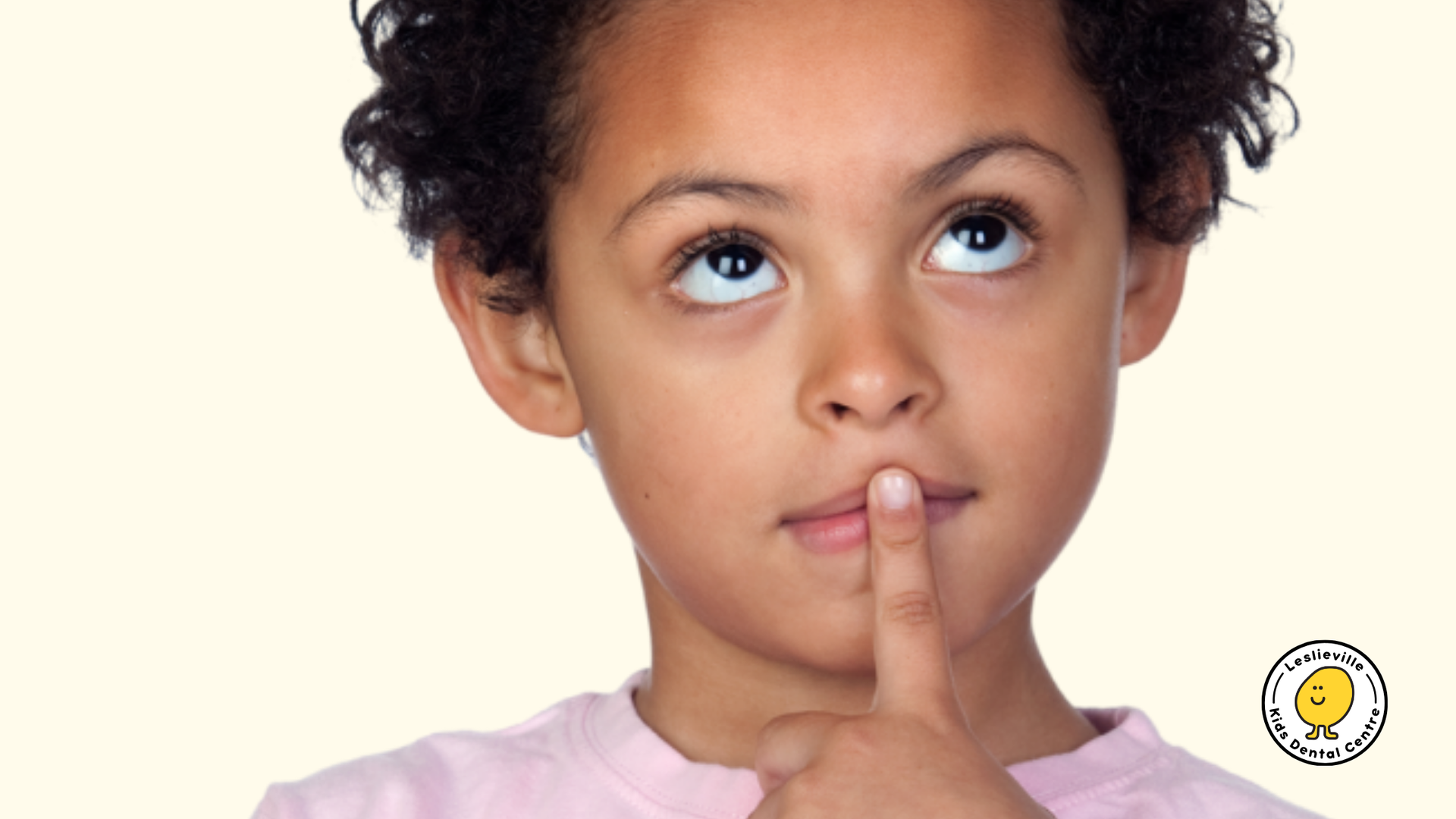 Child with curly hair looks upwards, finger to lips, thoughtful expression.