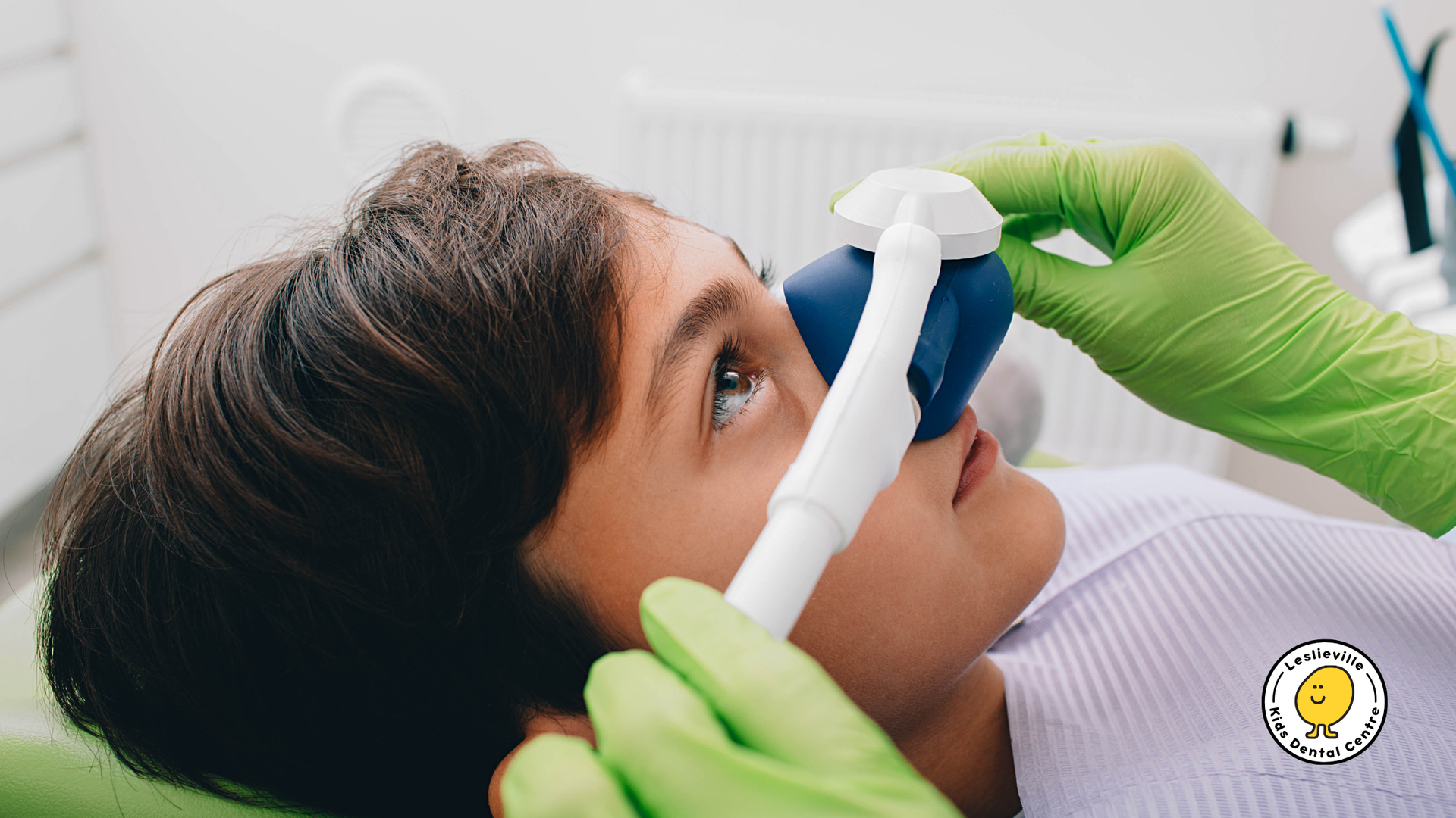 A person in a dental chair has a blue nasal mask for sedation placed on their nose by a practitioner in green gloves.