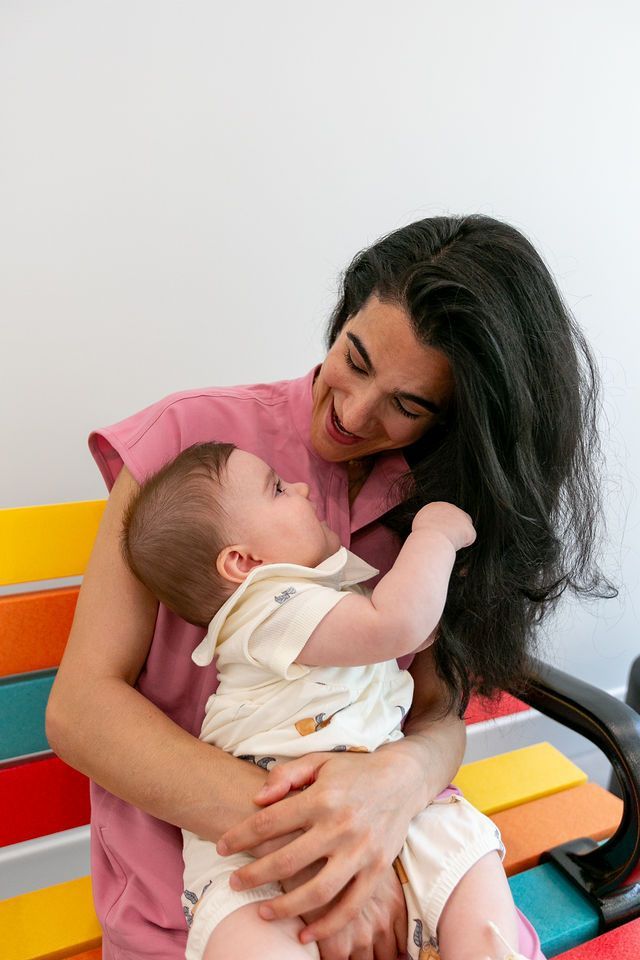 A person in a pink shirt smiling while holding a baby on a multicolored bench against a white wall.