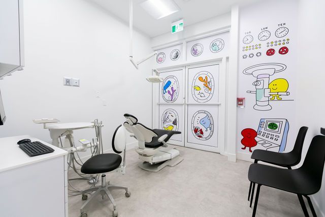 A child-friendly dental exam room with a reclining chair, equipment, and colorful wall murals of cartoon characters.