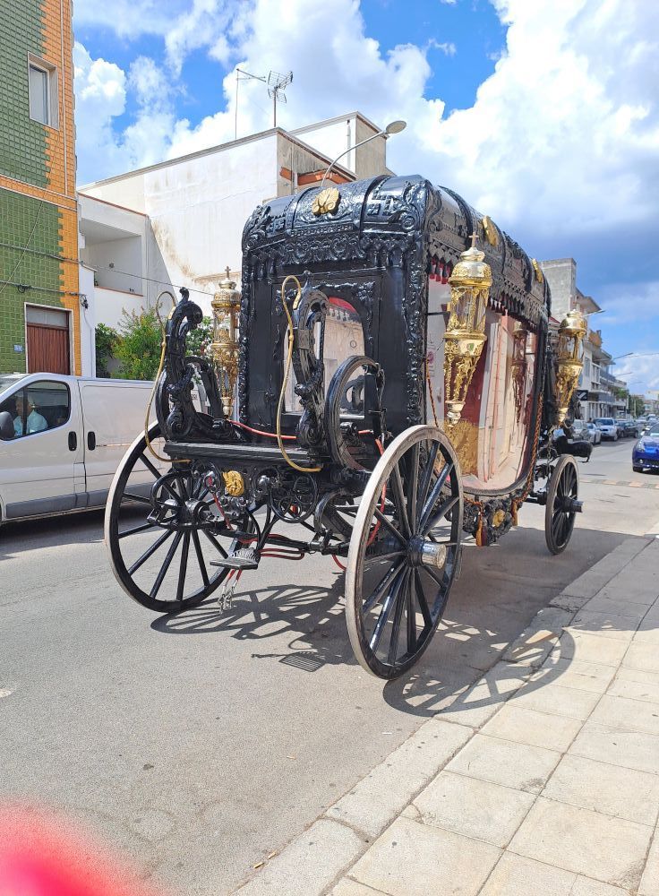 Una carrozza nera decorata, trainata da cavalli, parcheggiata in una strada soleggiata accanto a un furgone bianco.