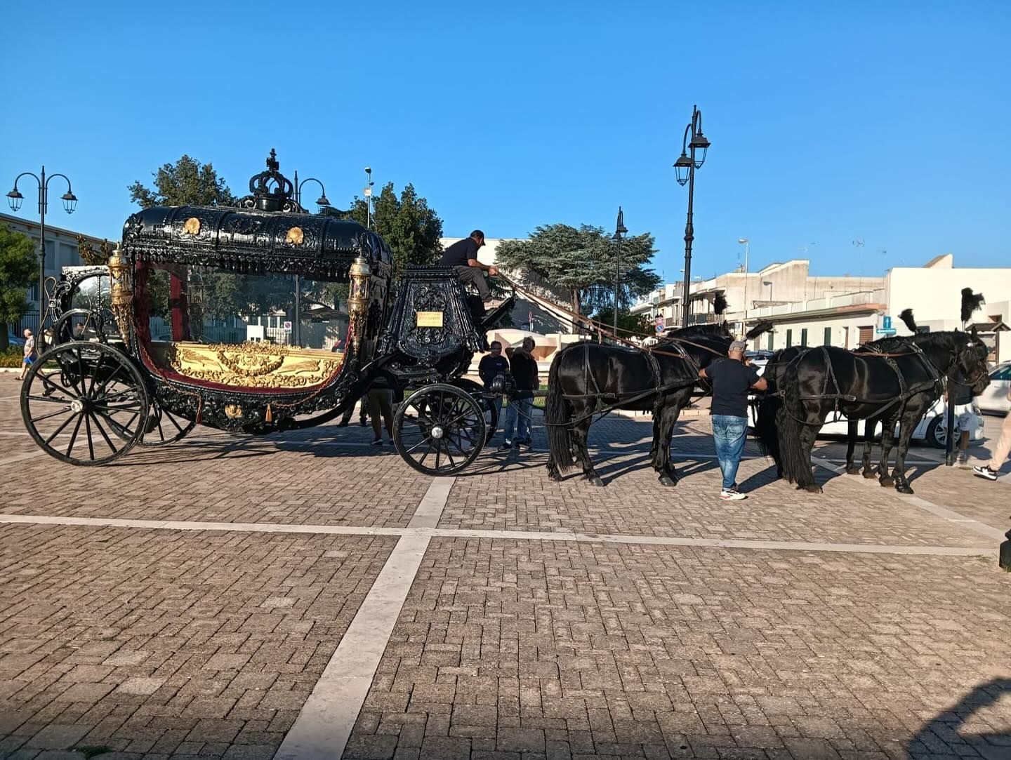Una carrozza nera trainata da cavalli su una piazza lastricata di ciottoli sotto un cielo azzurro brillante.