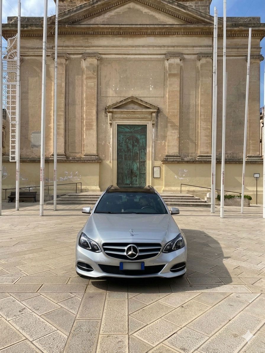 Una berlina Mercedes color argento parcheggiata davanti a un imponente edificio in pietra con colonne e un portale aperto.