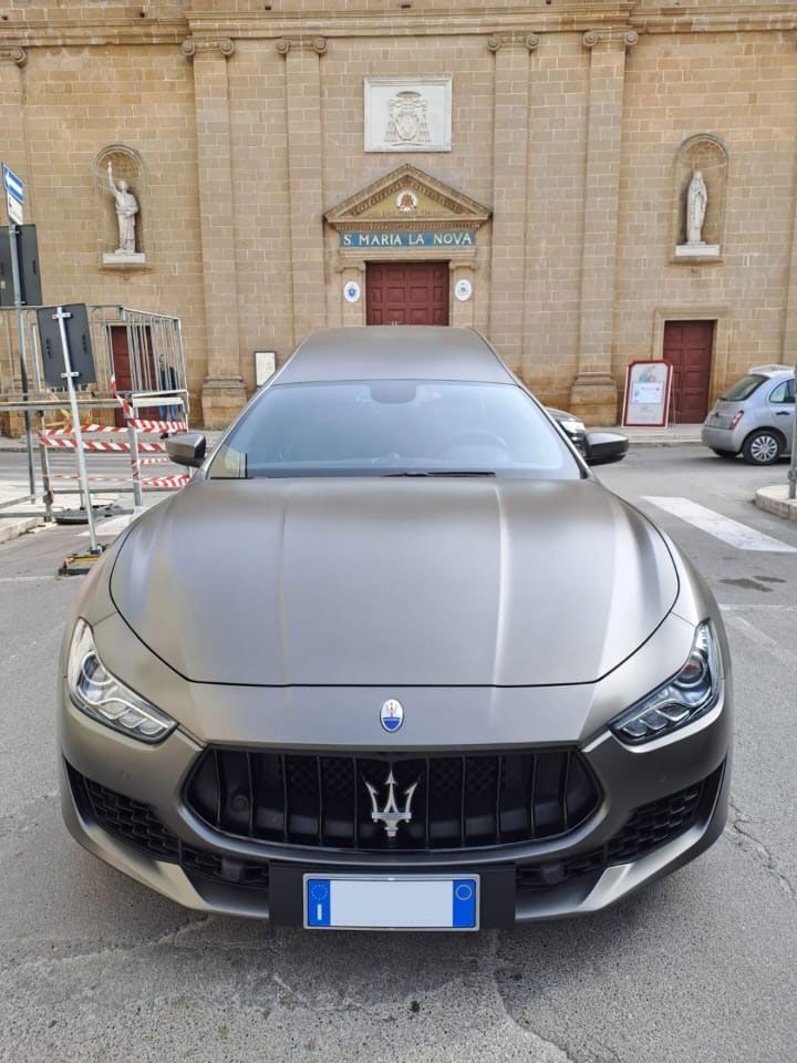 Vista frontale di una Maserati grigia parcheggiata in una strada cittadina di fronte a un edificio in mattoni.