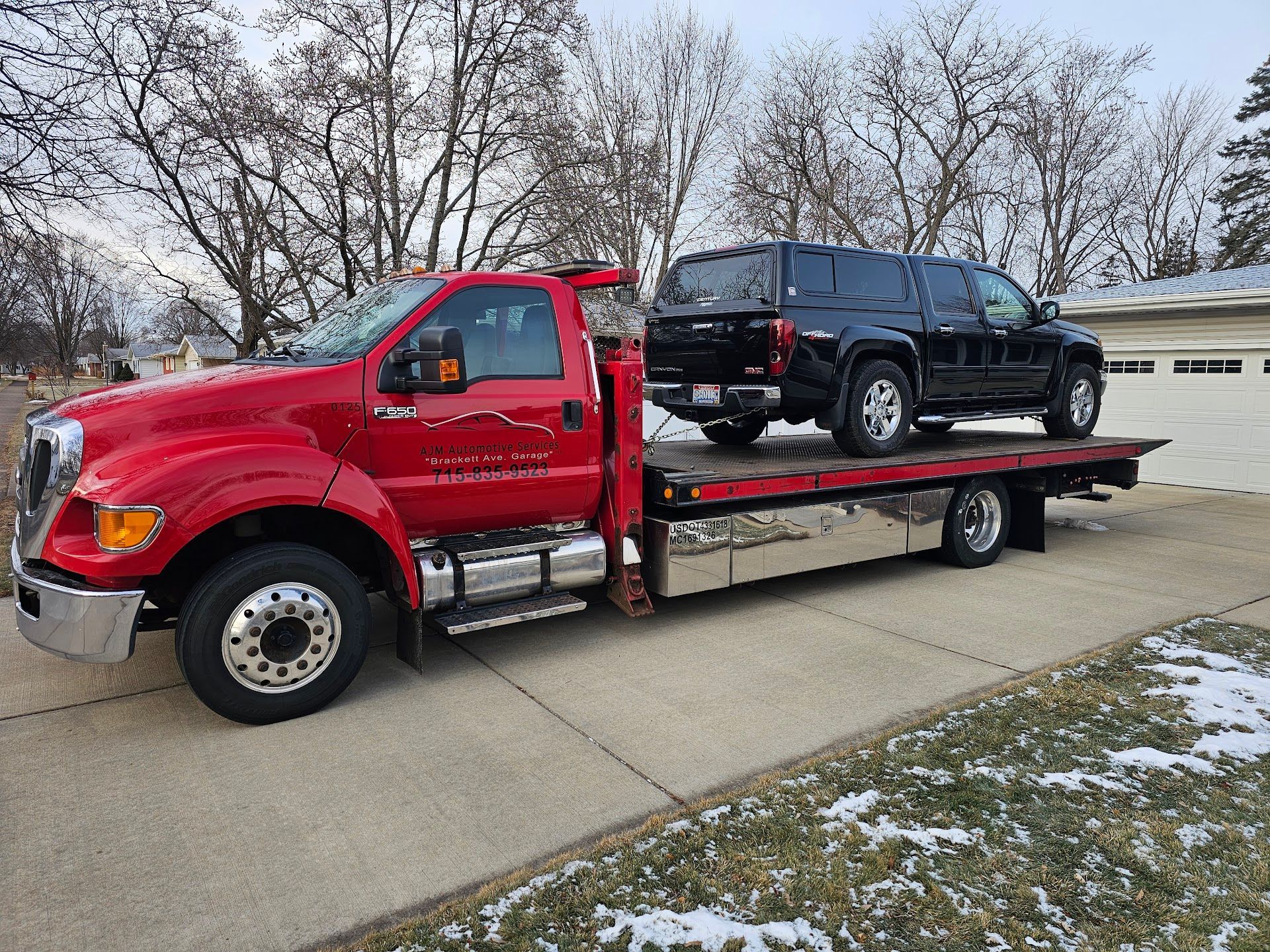 Red tow truck carrying a black SUV on a flatbed trailer in a residential driveway with snow on the ground  | AJM Automotive Services LLC