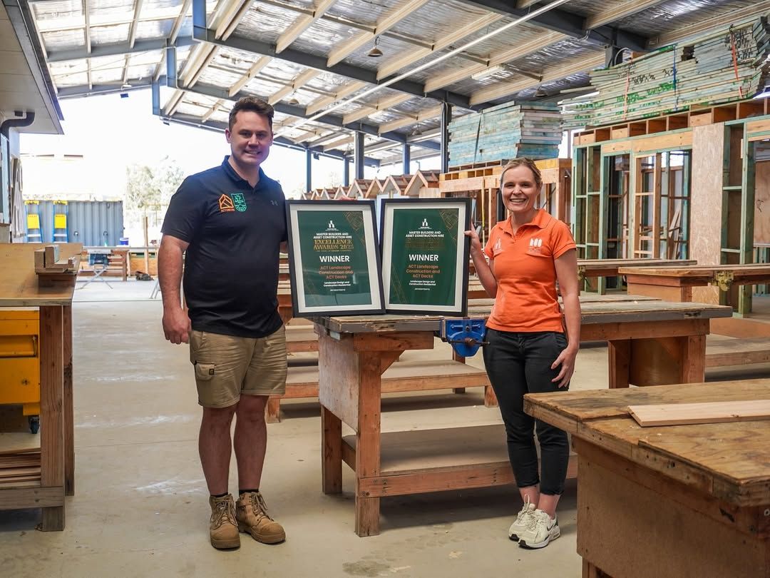 Two people standing in a workshop, holding two framed awards between them in front of a wooden workbench.