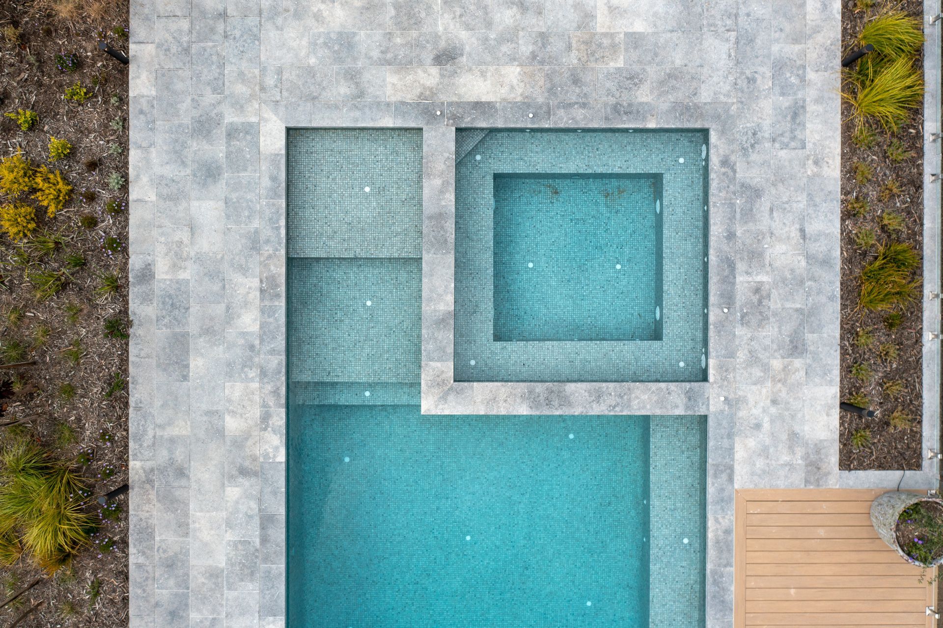 An aerial view of a turquoise swimming pool with an integrated spa, surrounded by grey stone pavers and garden beds.