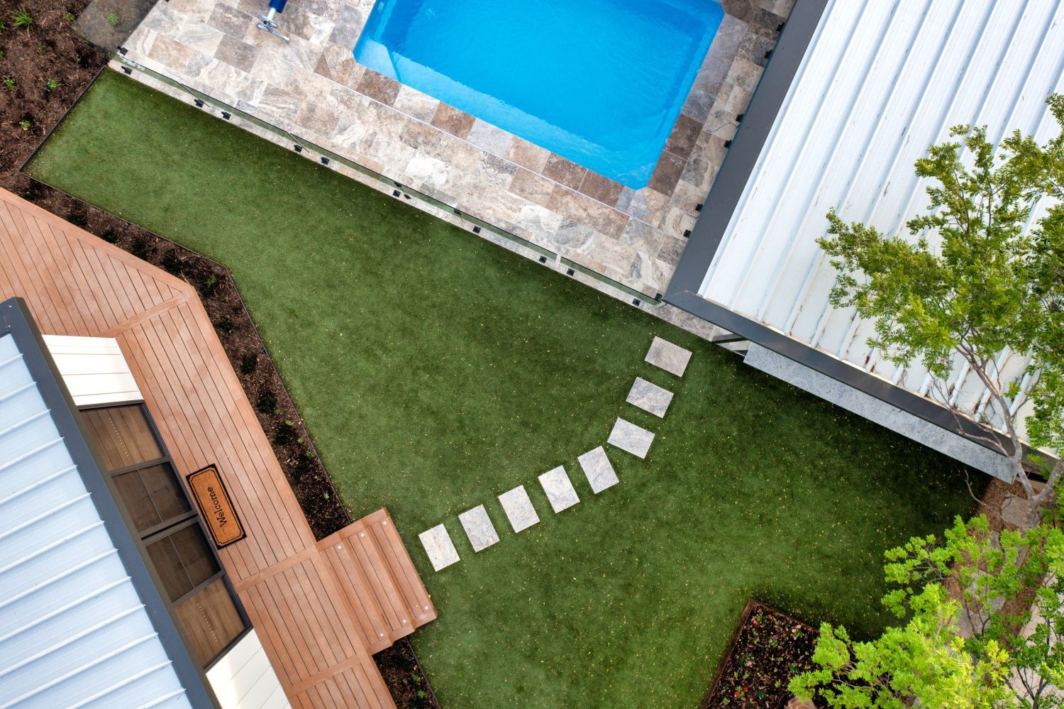 A top-down view of a backyard with a blue rectangular pool, green turf, a wooden deck, and a stone stepping stone path.