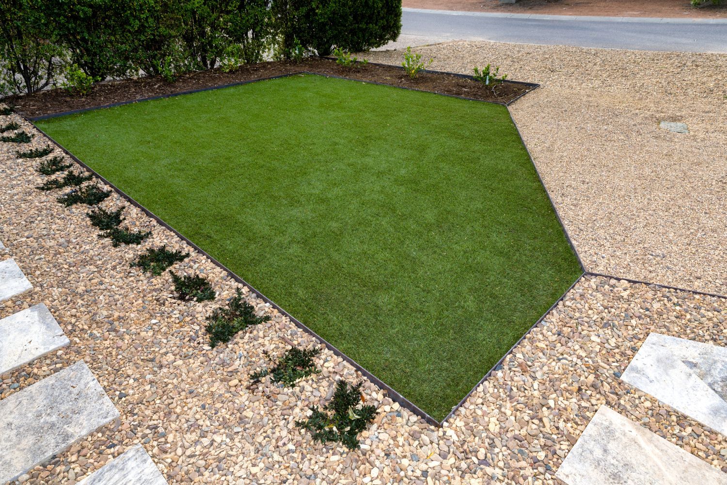 A geometric patch of green lawn bordered by beige gravel and stepping stones, with a small garden bed along the back.