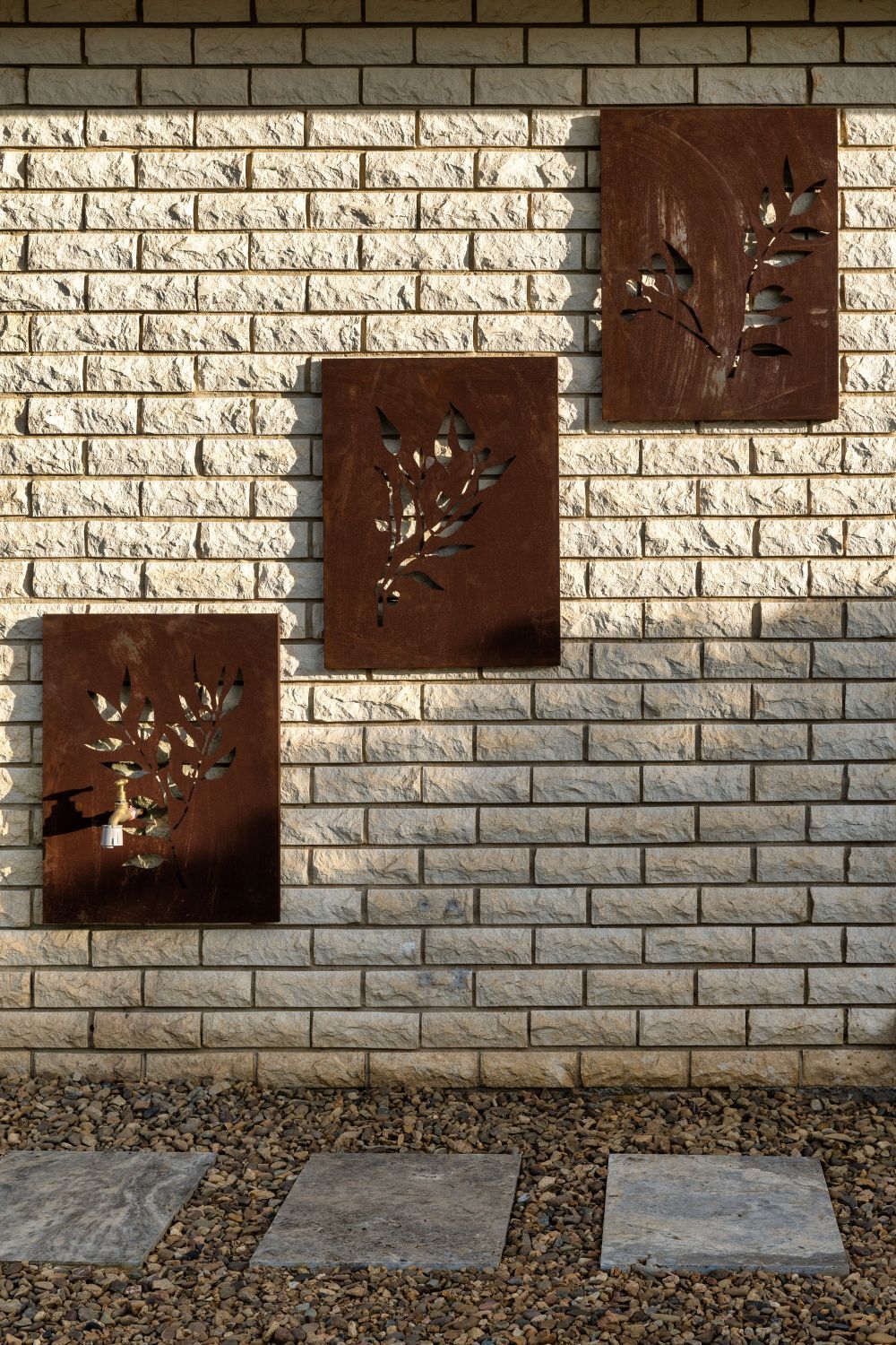 Three rectangular metal art panels with nature-themed cutouts mounted on a light-colored brick wall above stone pavers.