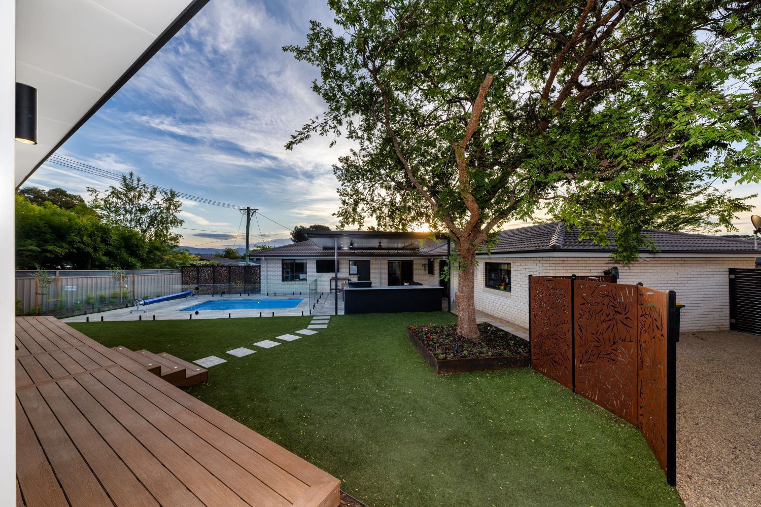 A backyard scene with a wooden deck, artificial lawn, swimming pool, mature tree, and decorative metal privacy panels.
