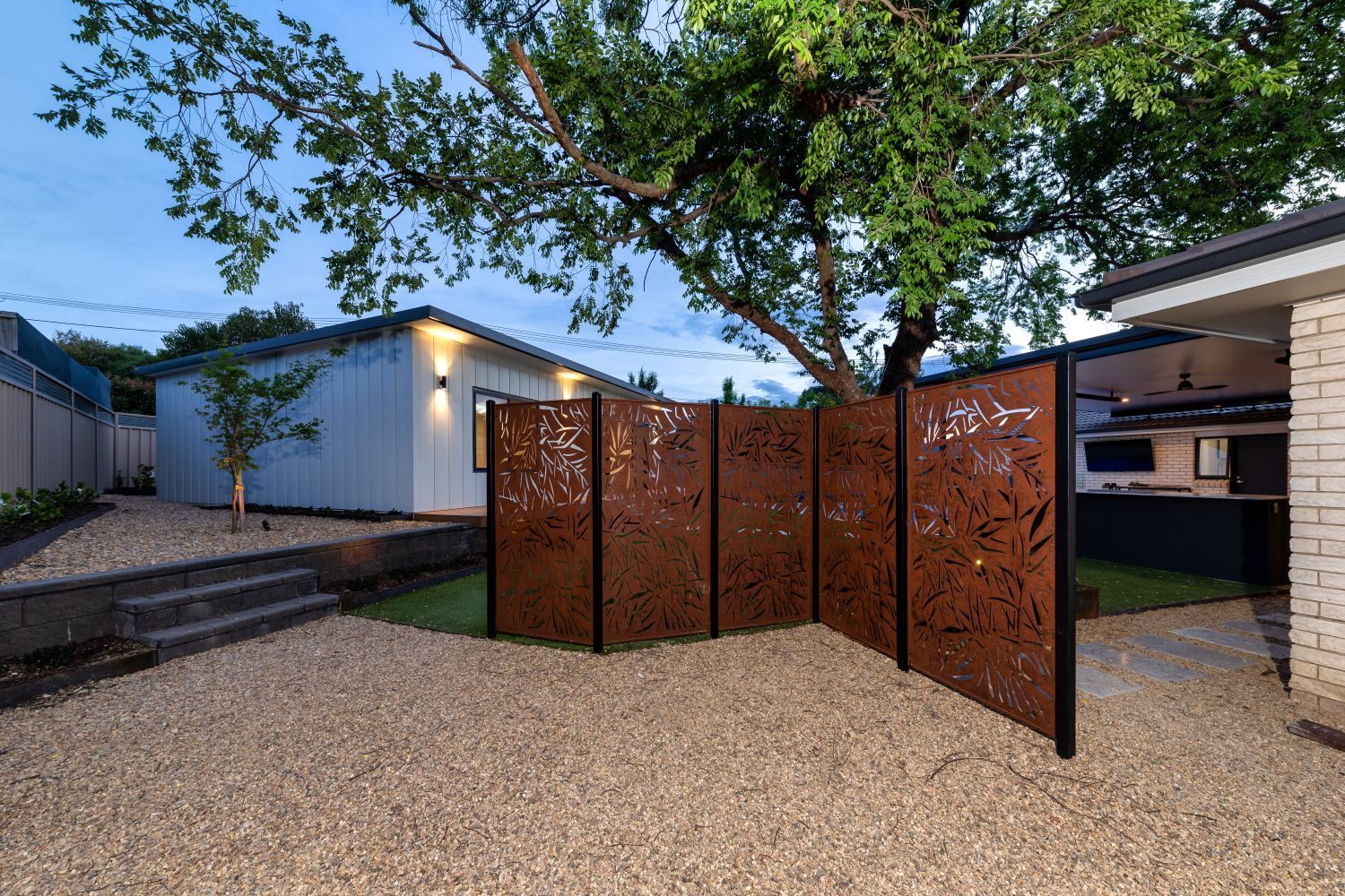 A decorative metal privacy screen stands in a gravel-covered backyard at dusk, separating a patio from the garden.