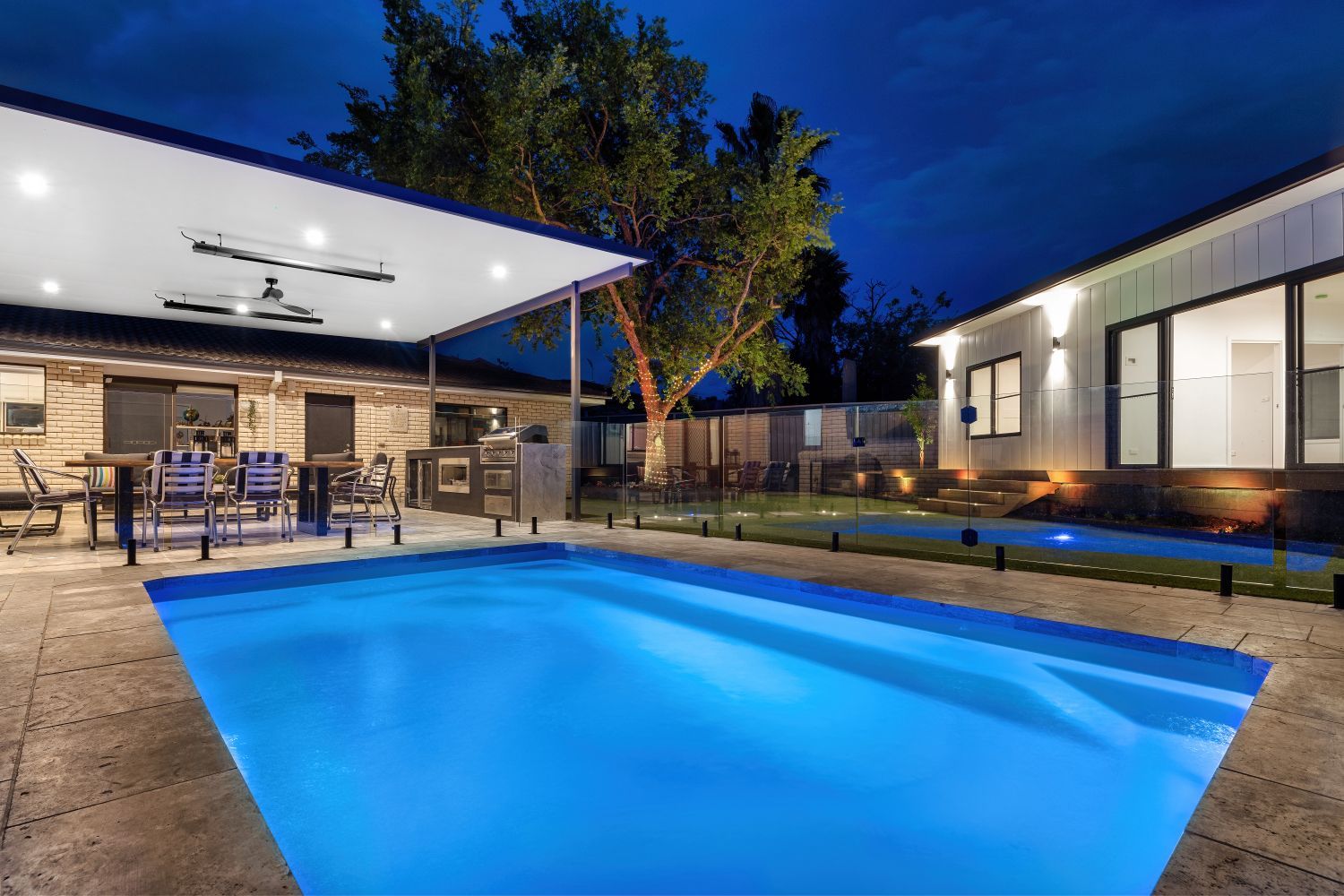 A brightly illuminated swimming pool sits before a modern patio and house at twilight.