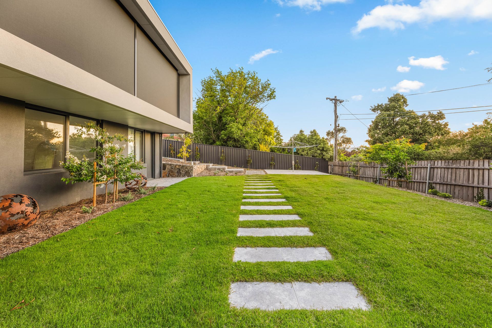 A modern house with a gray exterior overlooks a green lawn featuring a straight stone path, with trees and a fence beyond.