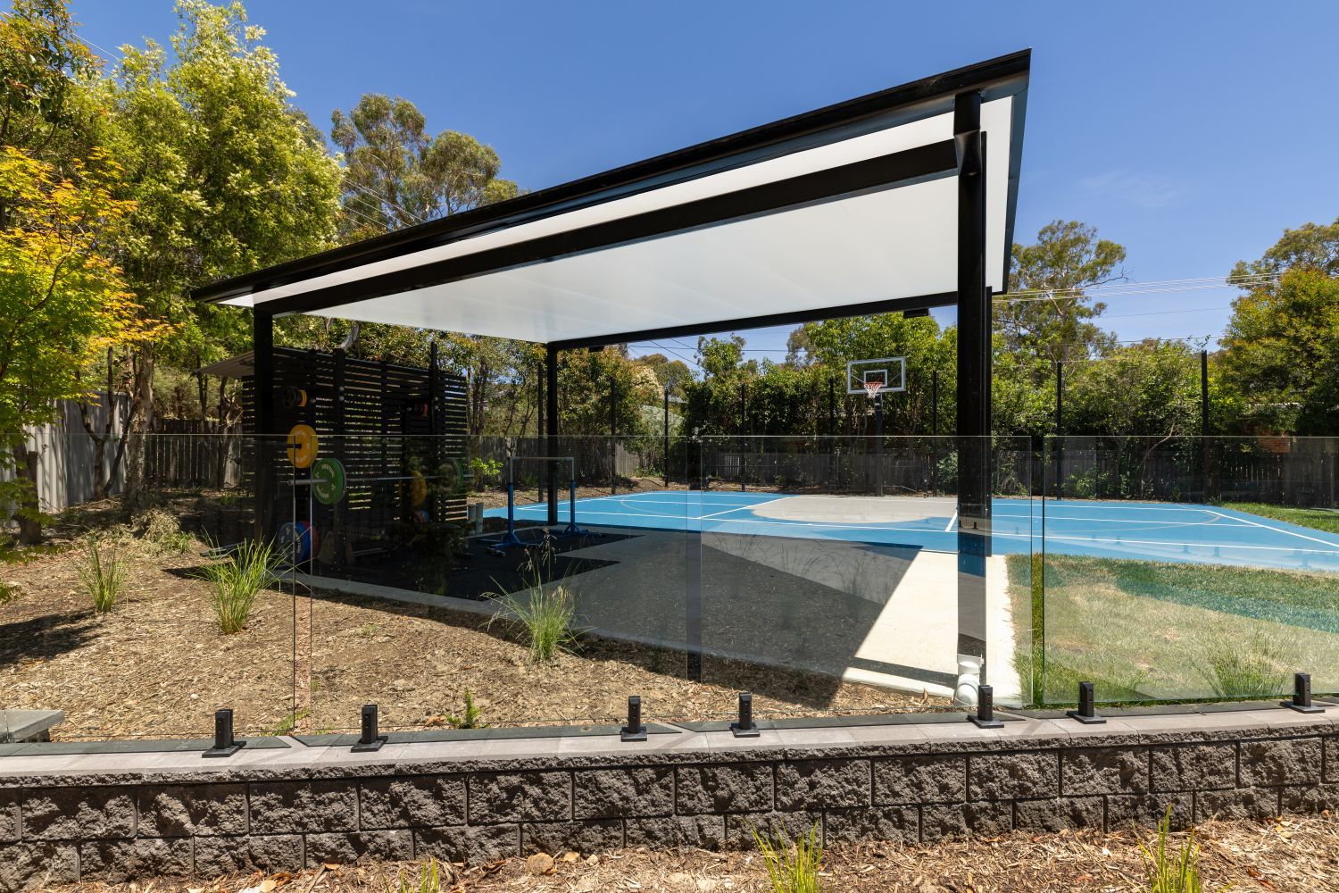 A covered outdoor patio with a black frame and white roof overlooking a blue basketball court in a sunny, wooded area.