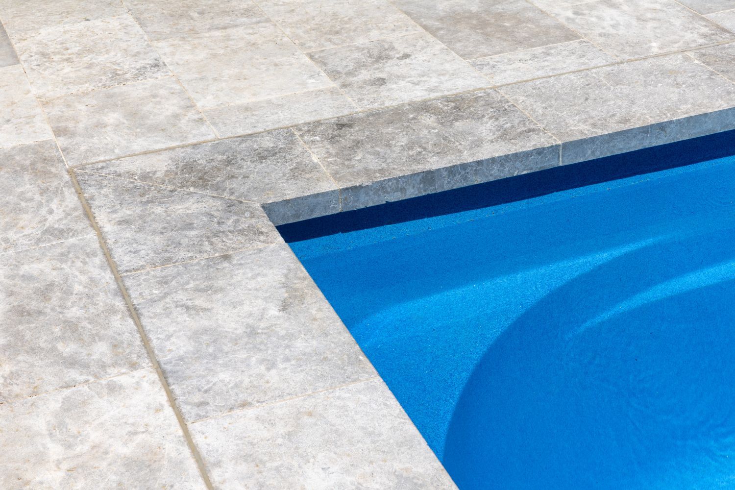 A bright blue swimming pool edge with textured light gray stone pavers.