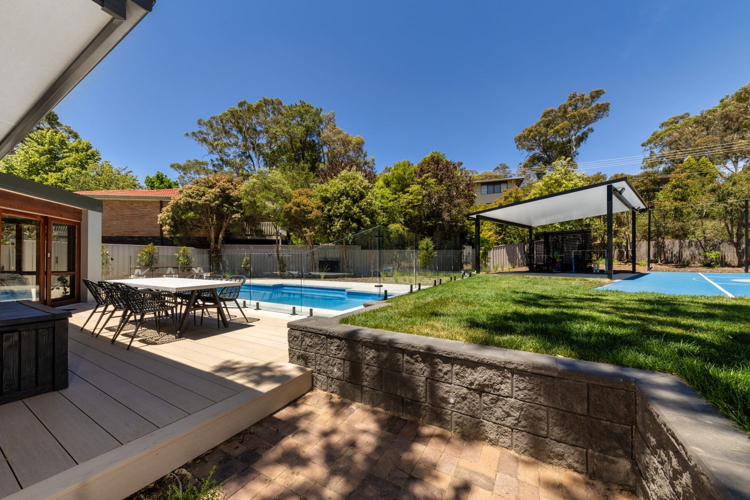 A backyard featuring a wooden deck with a table, a swimming pool, a grassy lawn, and a shaded outdoor seating area.