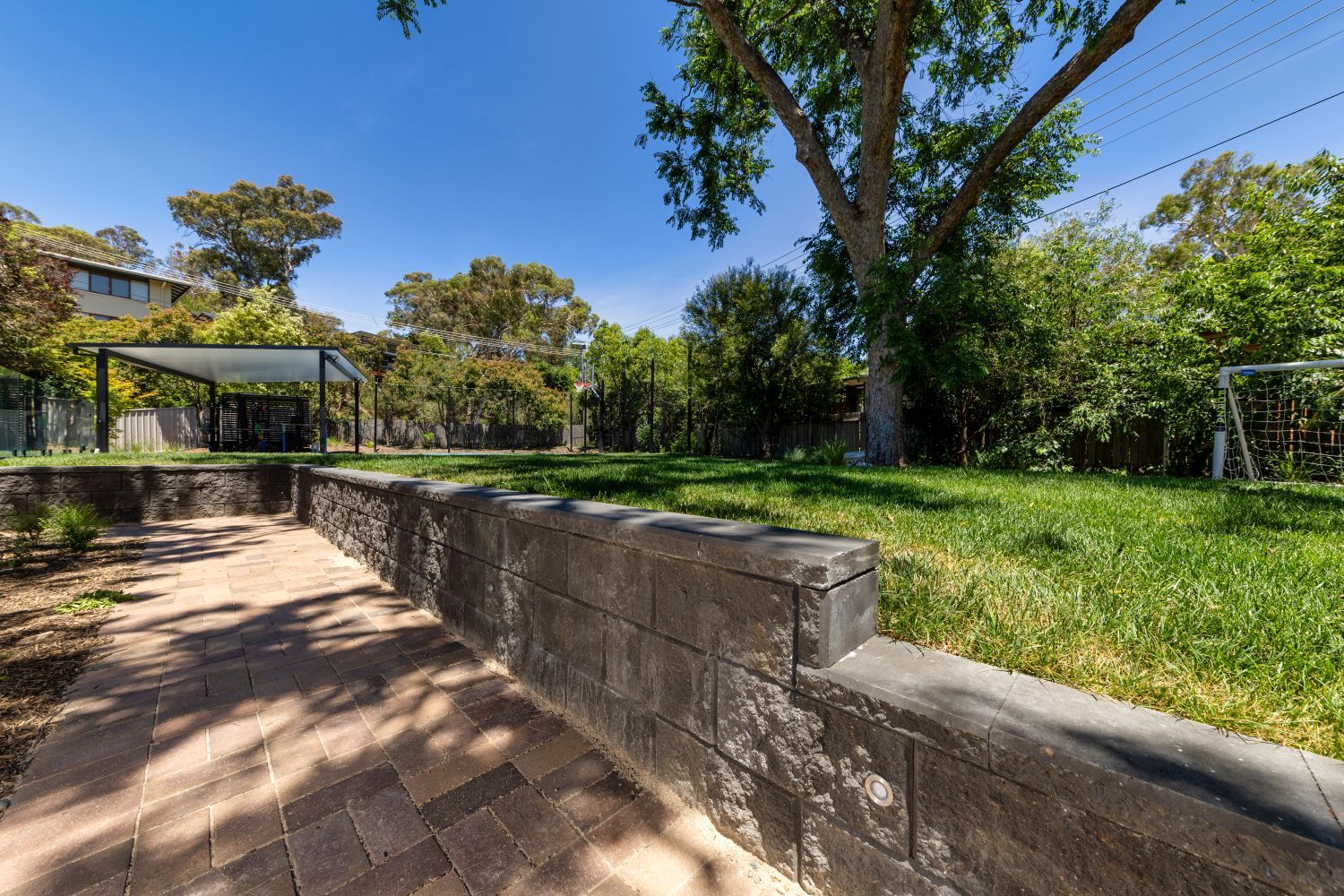 A backyard view featuring a paved patio, a stone retaining wall, and a grassy lawn under a clear blue sky.