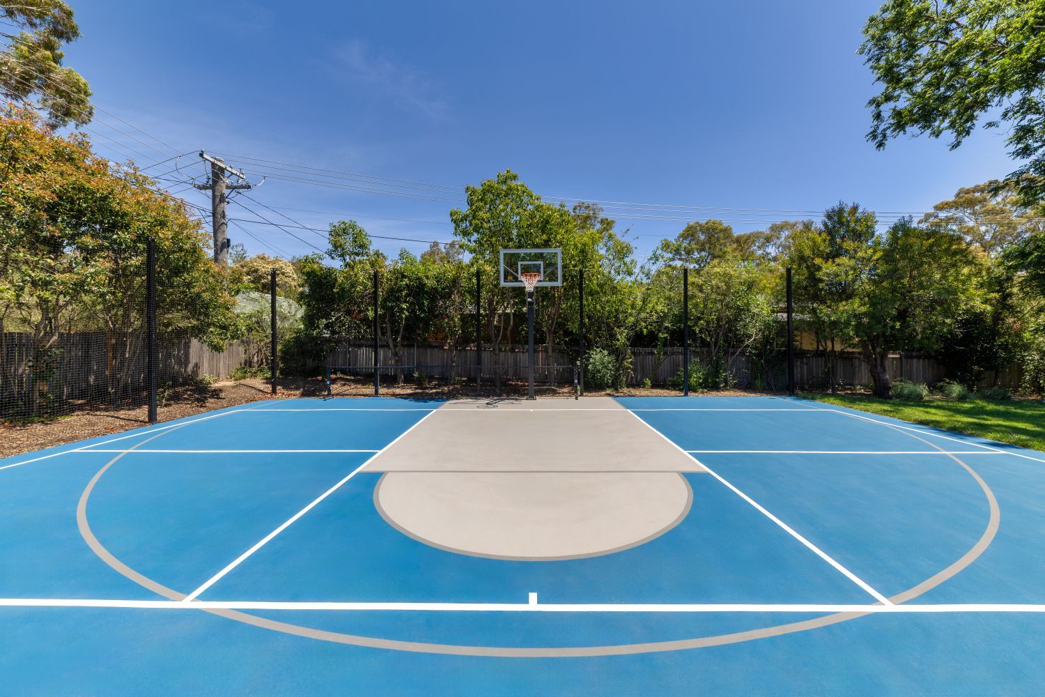 A blue and grey outdoor basketball court surrounded by trees under a clear blue sky.