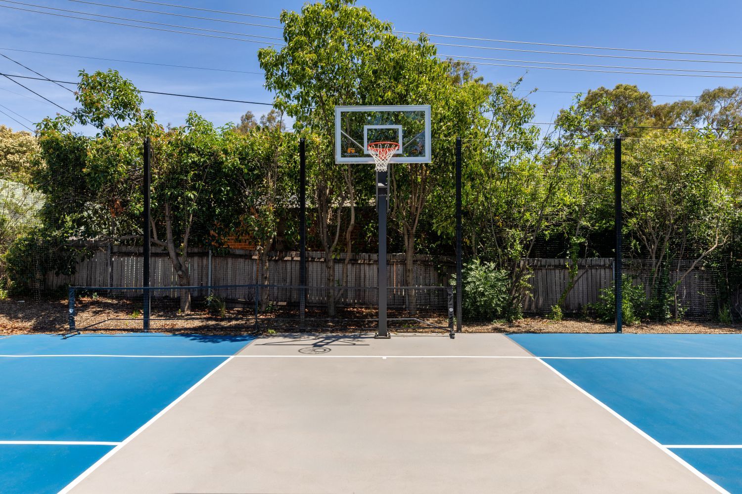 A basketball hoop centered on a blue and grey outdoor court, backed by a wooden fence and lush green trees.