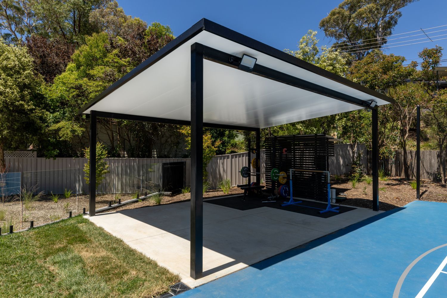 An outdoor gym with fitness equipment under a black metal-framed roof, situated next to a blue sports court.