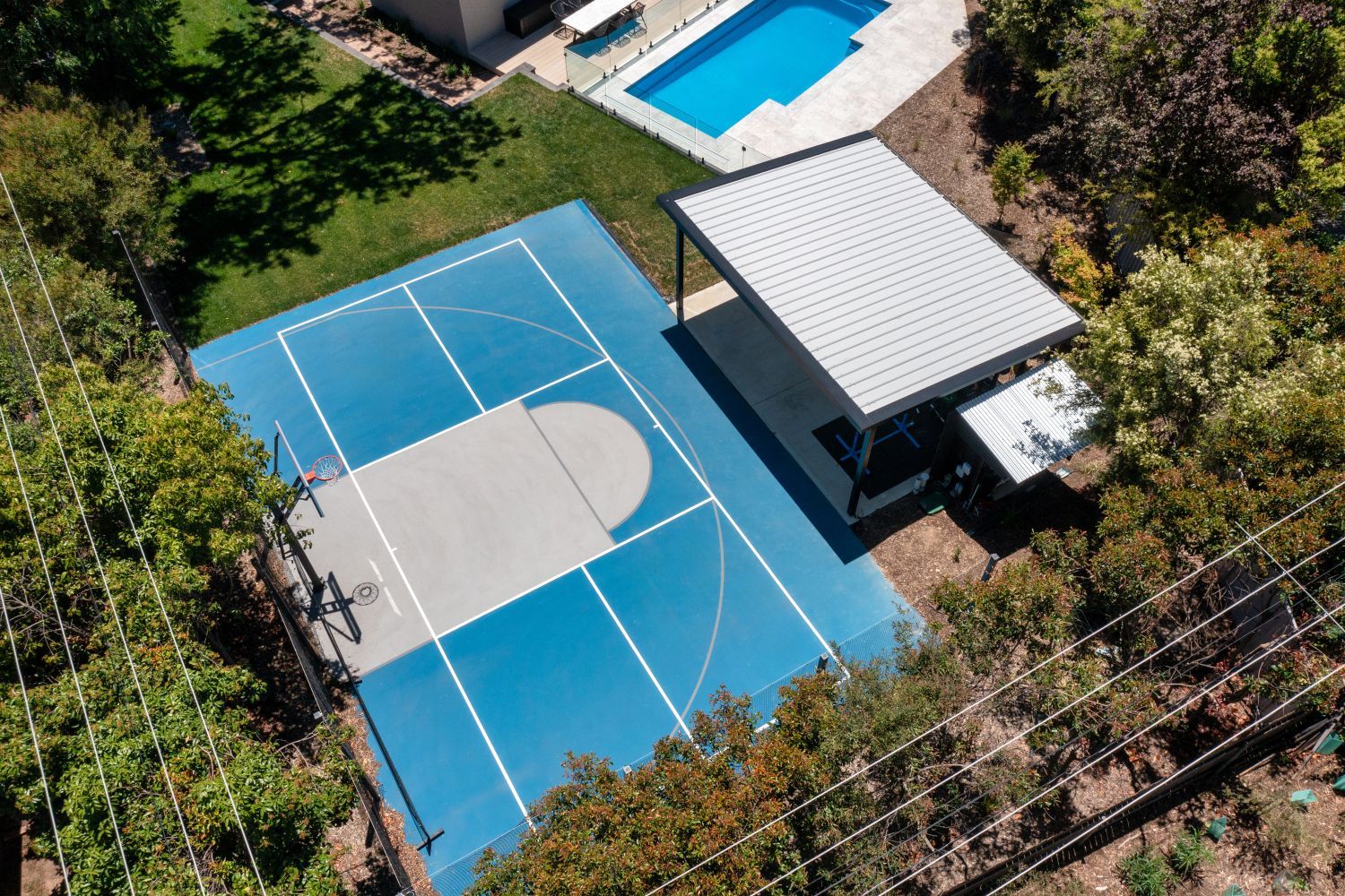 An aerial view of a blue and grey basketball court next to a small pavilion and a rectangular swimming pool.