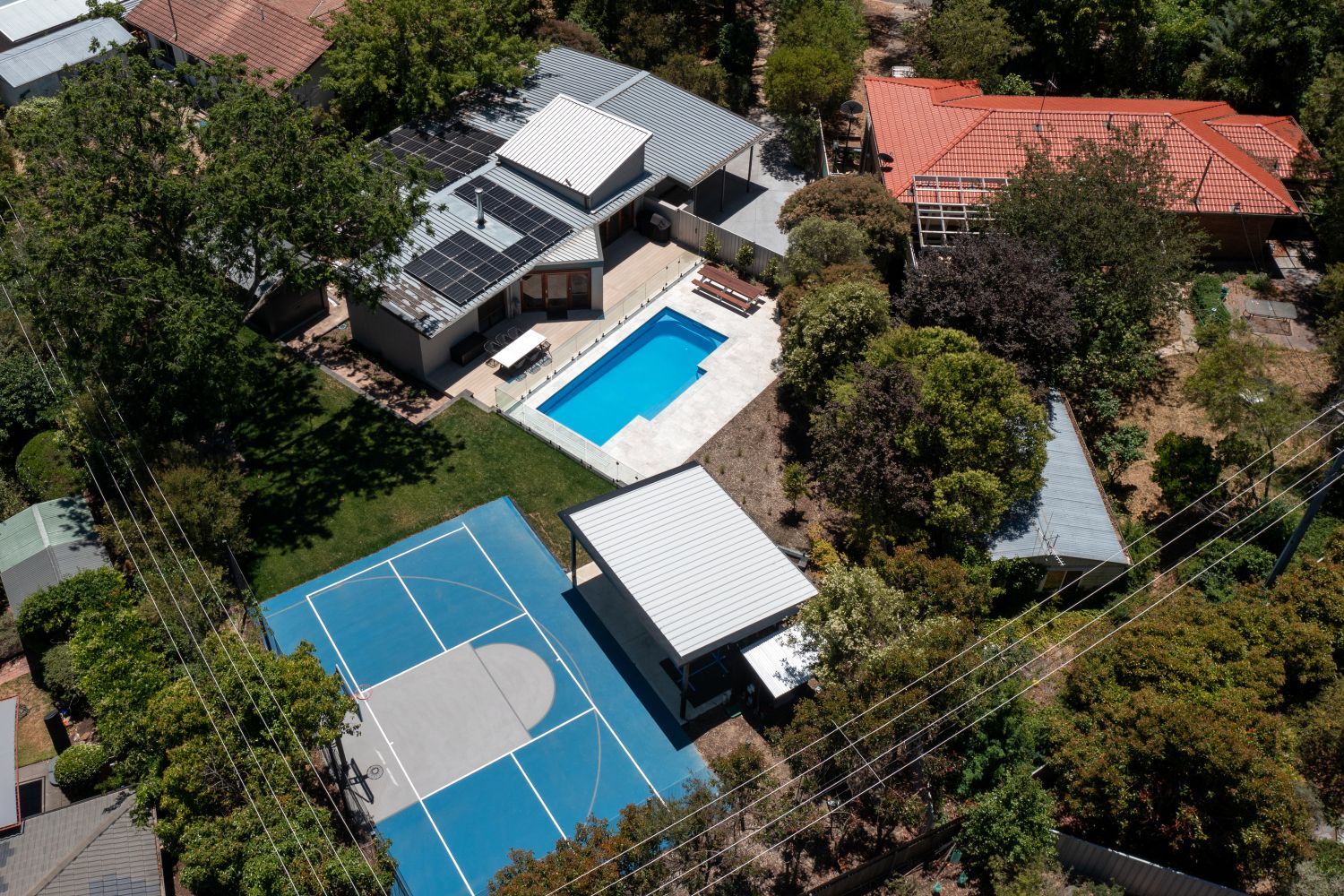 An aerial view shows a house with solar panels, a blue swimming pool, a small white building, and a backyard basketball court.