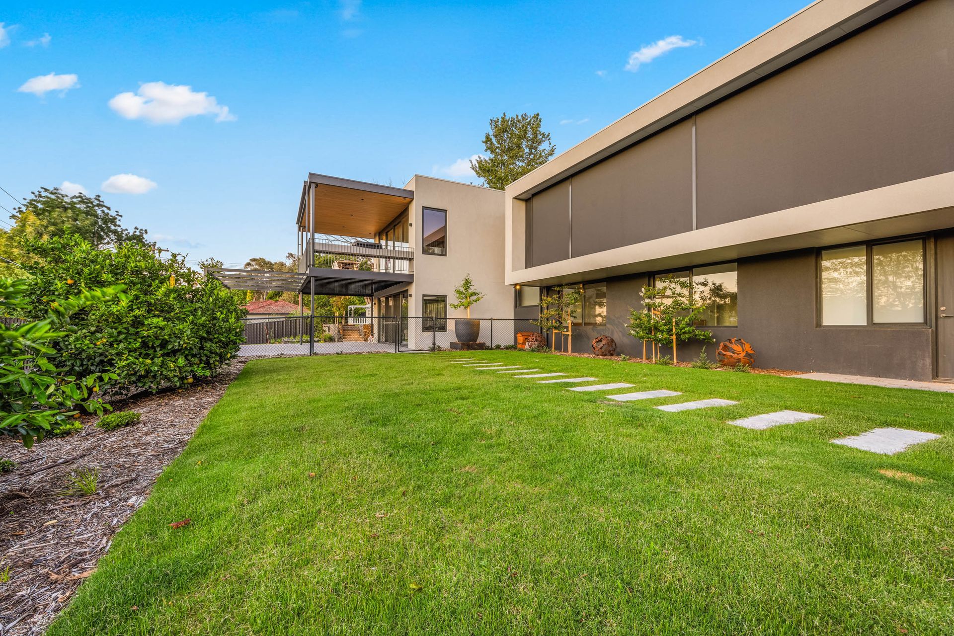 Modern two-story house with a flat roof, large windows, and an upper deck overlooking a green lawn with stone pavers.