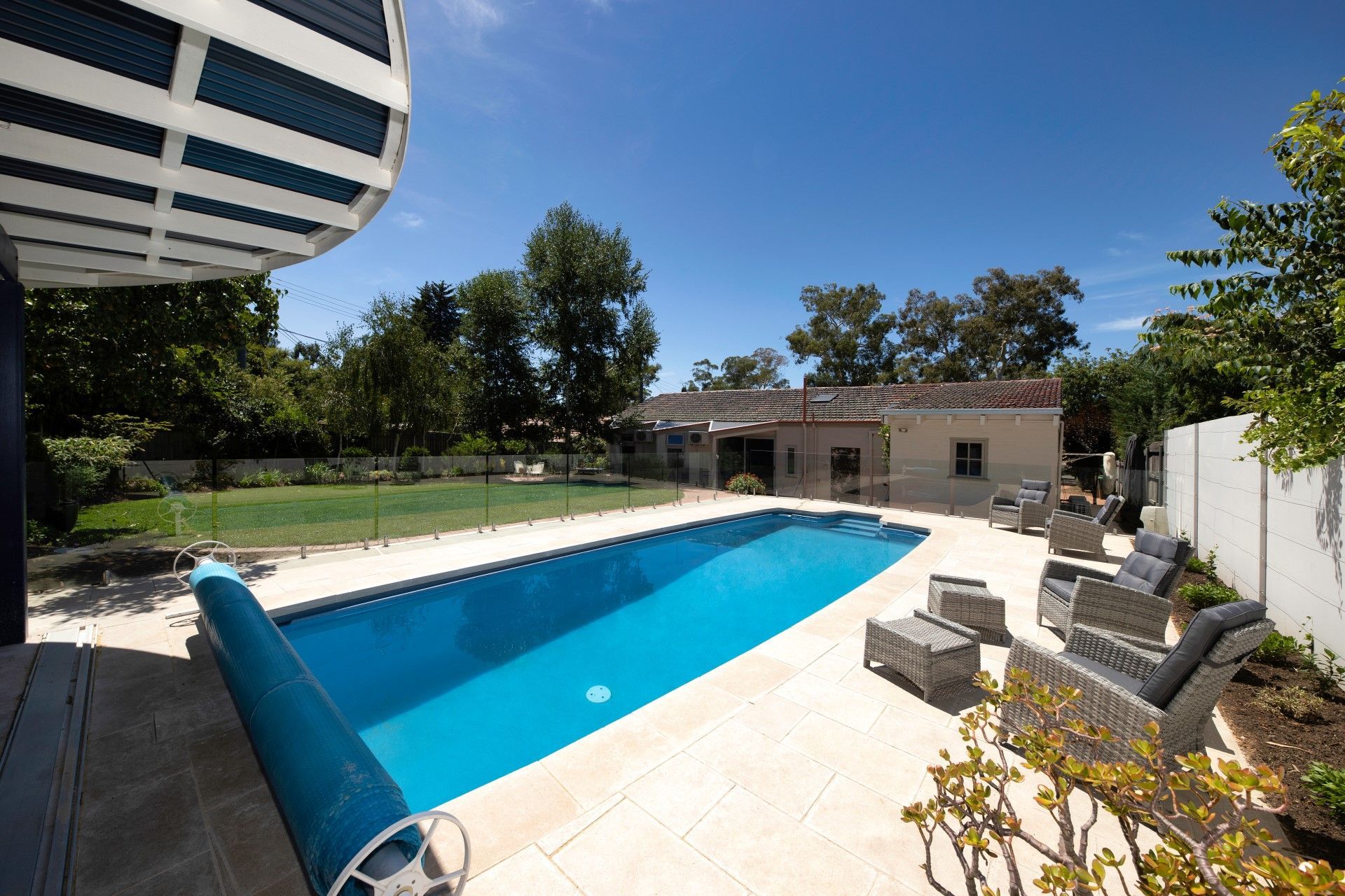 A bright blue rectangular swimming pool with a partially rolled-up cover on a stone patio, adjacent to a grassy backyard.