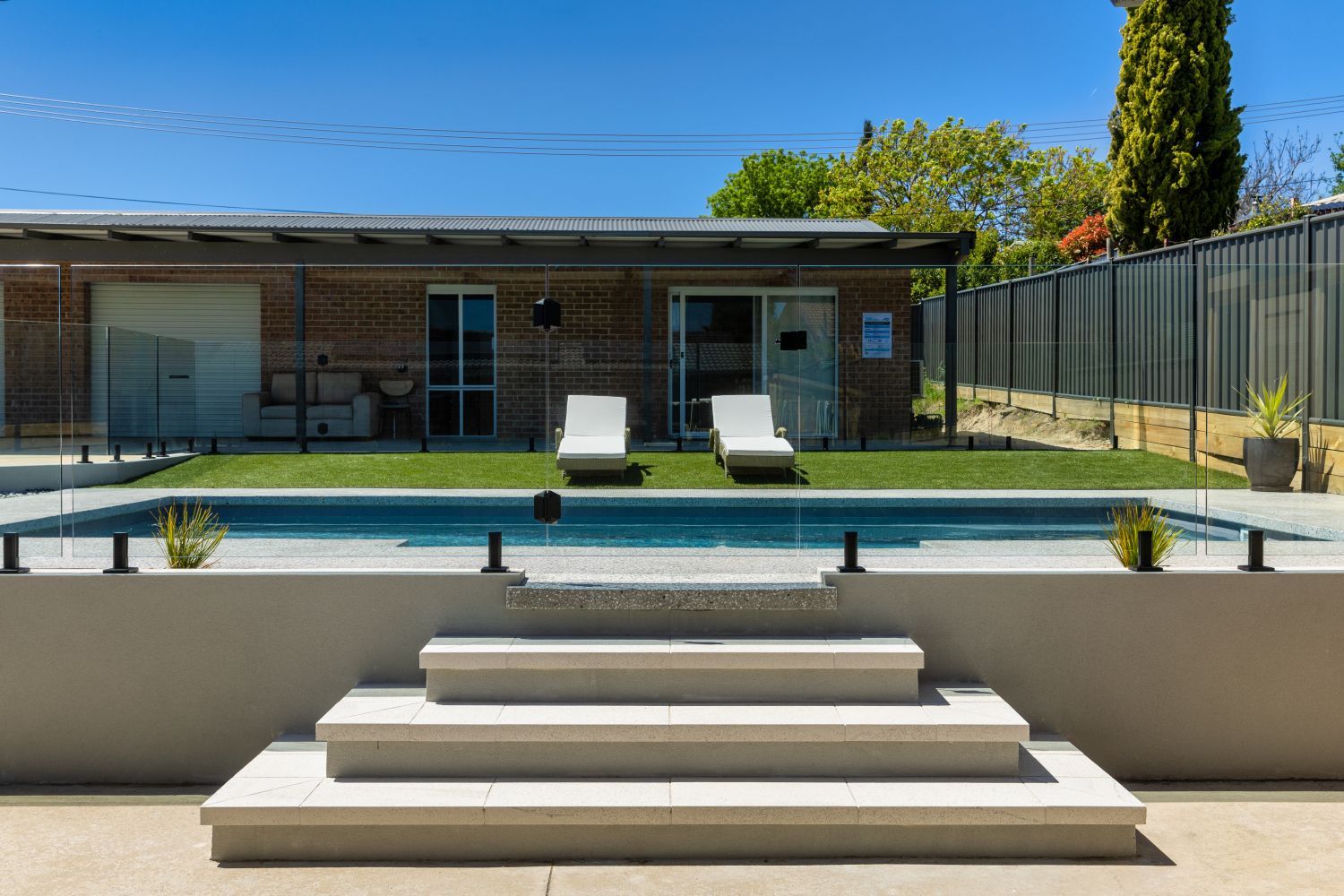 A swimming pool with three wide stone steps leading up to a grassy backyard and a modern single-story brick house.