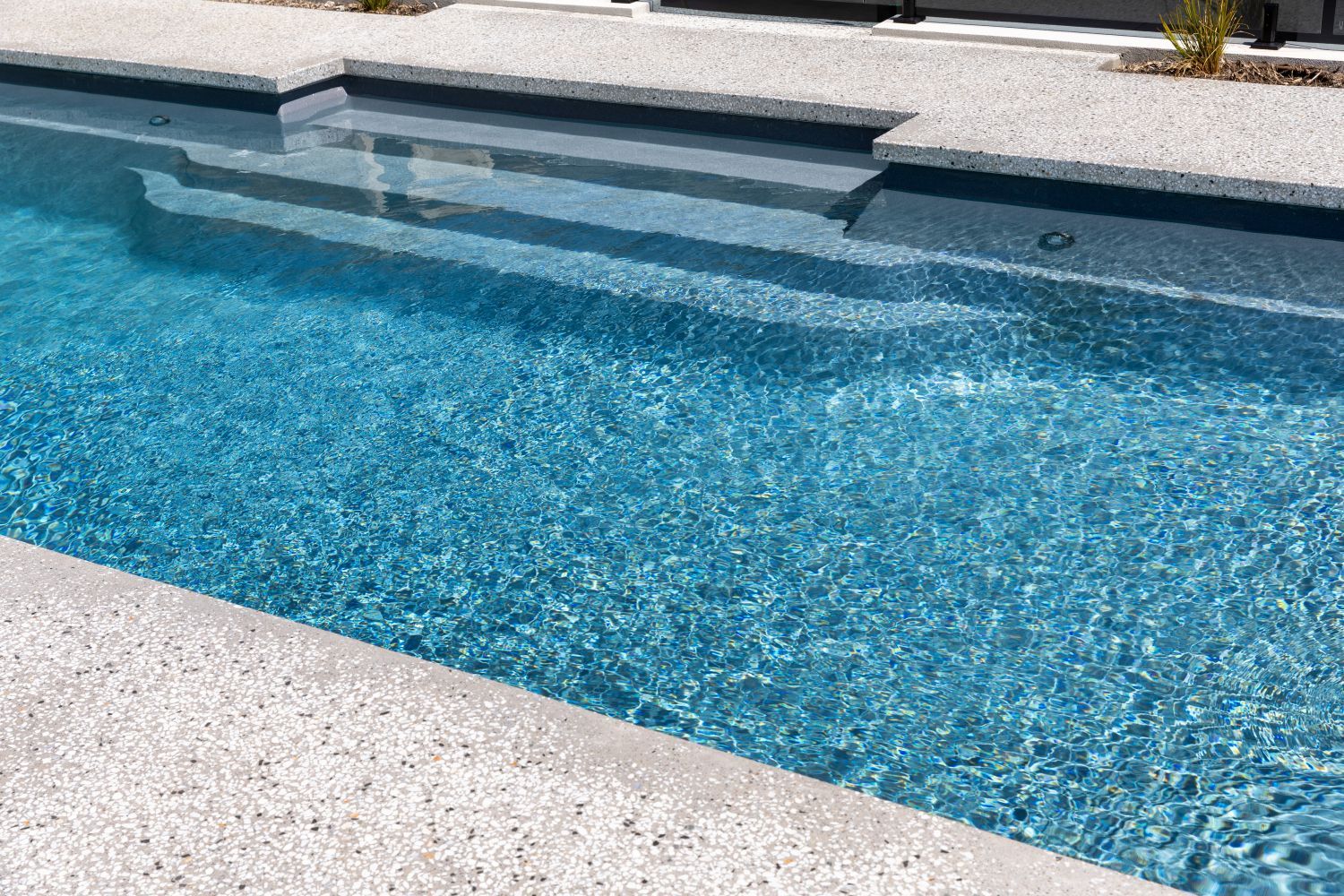 A bright blue swimming pool with steps leading into the water, bordered by a textured, light-colored concrete deck.