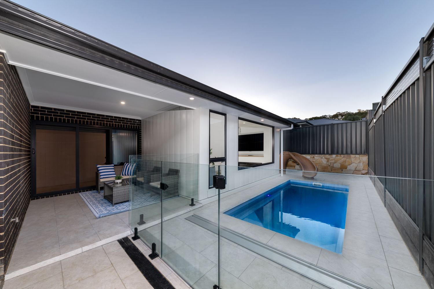 A modern patio and outdoor living space with a swimming pool, glass fencing, and seating area under a covered roof.