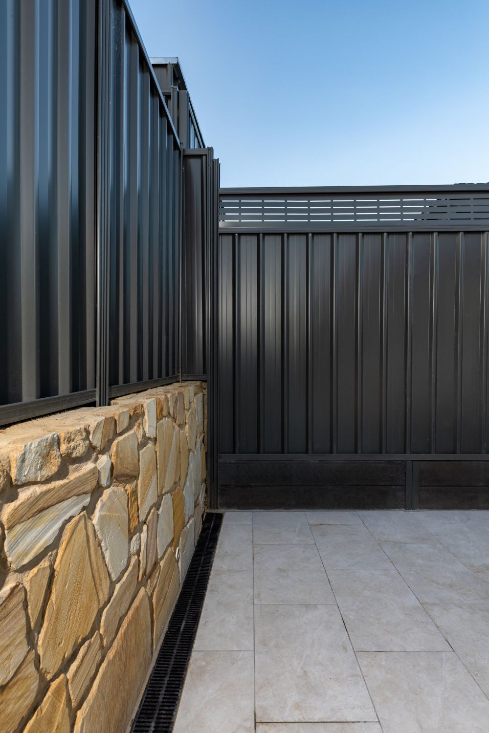A corner patio space featuring a stone wall, charcoal-colored metal fencing, and light gray tiled flooring.