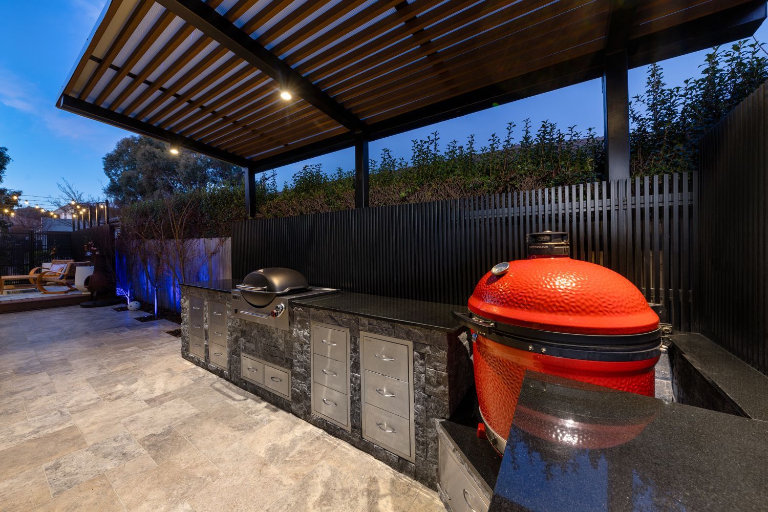 Outdoor kitchen with a built-in grill and a large, red ceramic kamado-style grill under a modern wooden pergola.