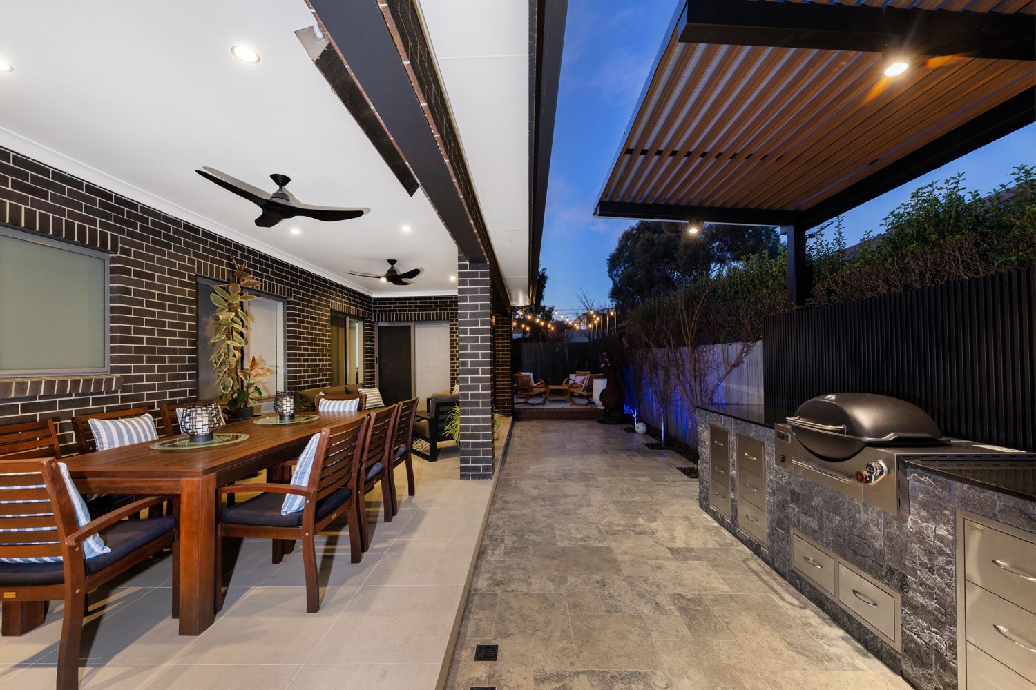 A modern outdoor patio at dusk with a wooden dining table, black brick walls, a grilling station, and a metal pergola.