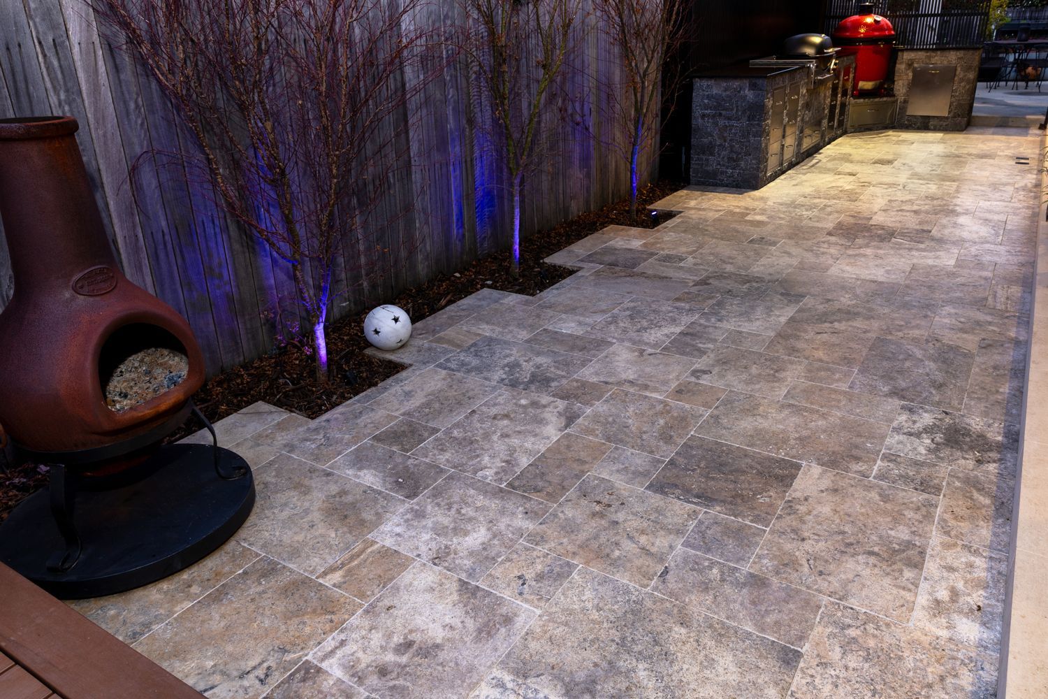 A patio paved with rectangular stone tiles, featuring a clay chiminea, small trees with purple lighting, and a soccer ball.
