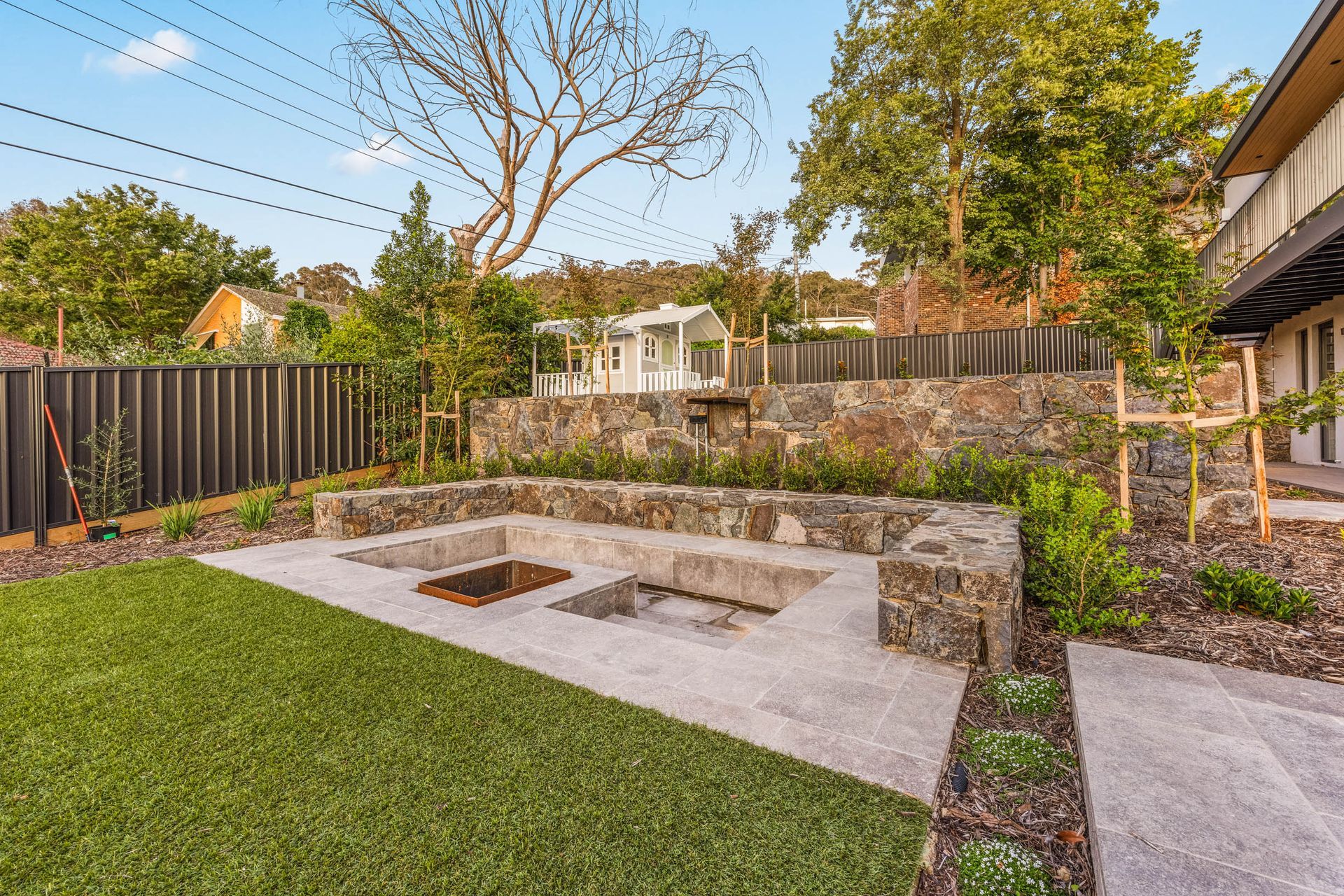 A sunken fire pit area with stone seating in a backyard featuring a green lawn, retaining walls, and surrounding trees.