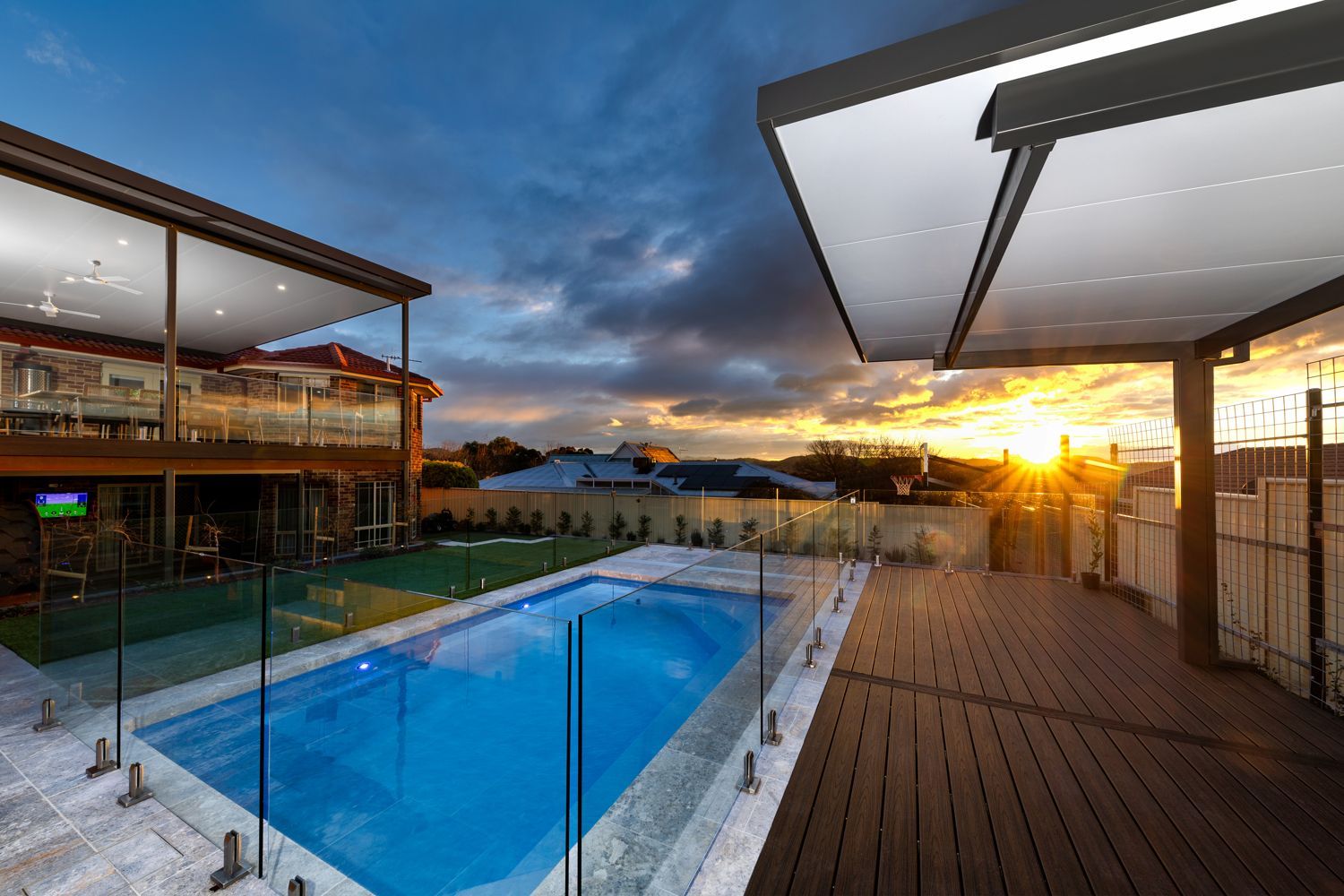 A swimming pool on a patio with a wooden deck and overhead canopy at sunset, seen from a house balcony.