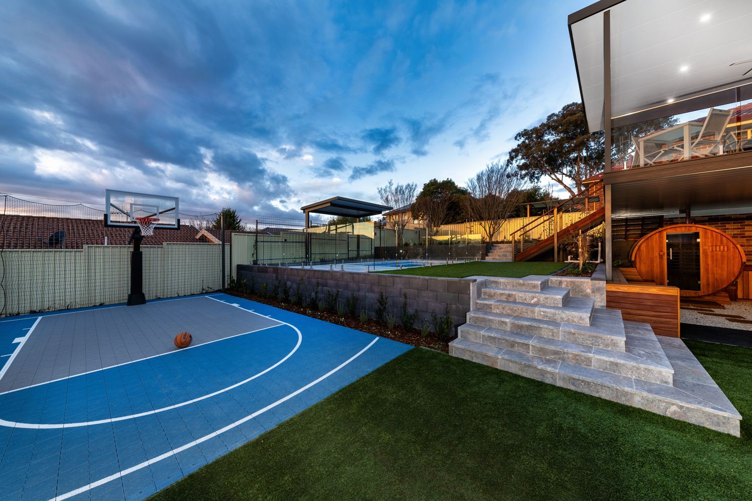 A backyard with a blue basketball court, green lawn, stone stairs, and a wooden barrel sauna under a deck at dusk.