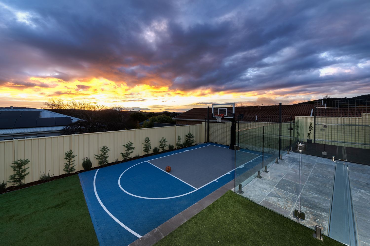 Backyard with a blue and gray basketball court next to a patio under a dramatic, colorful sunset sky.