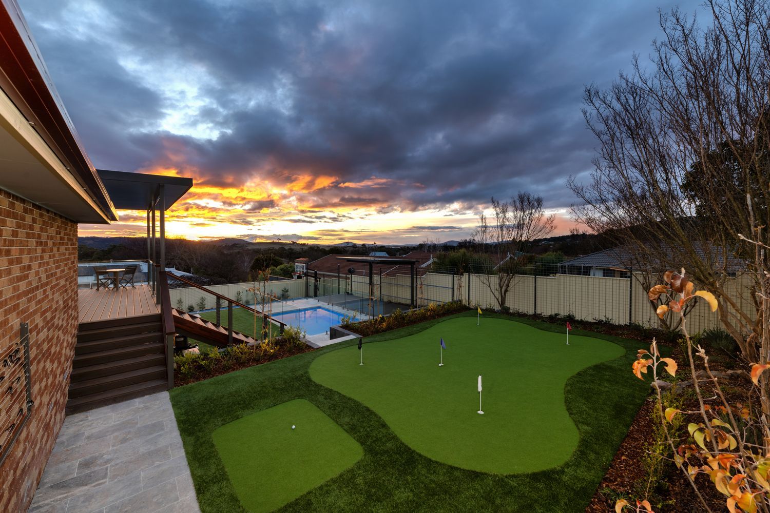 A backyard at dusk featuring a putting green, a small swimming pool, a wooden deck, and a vibrant sunset sky.