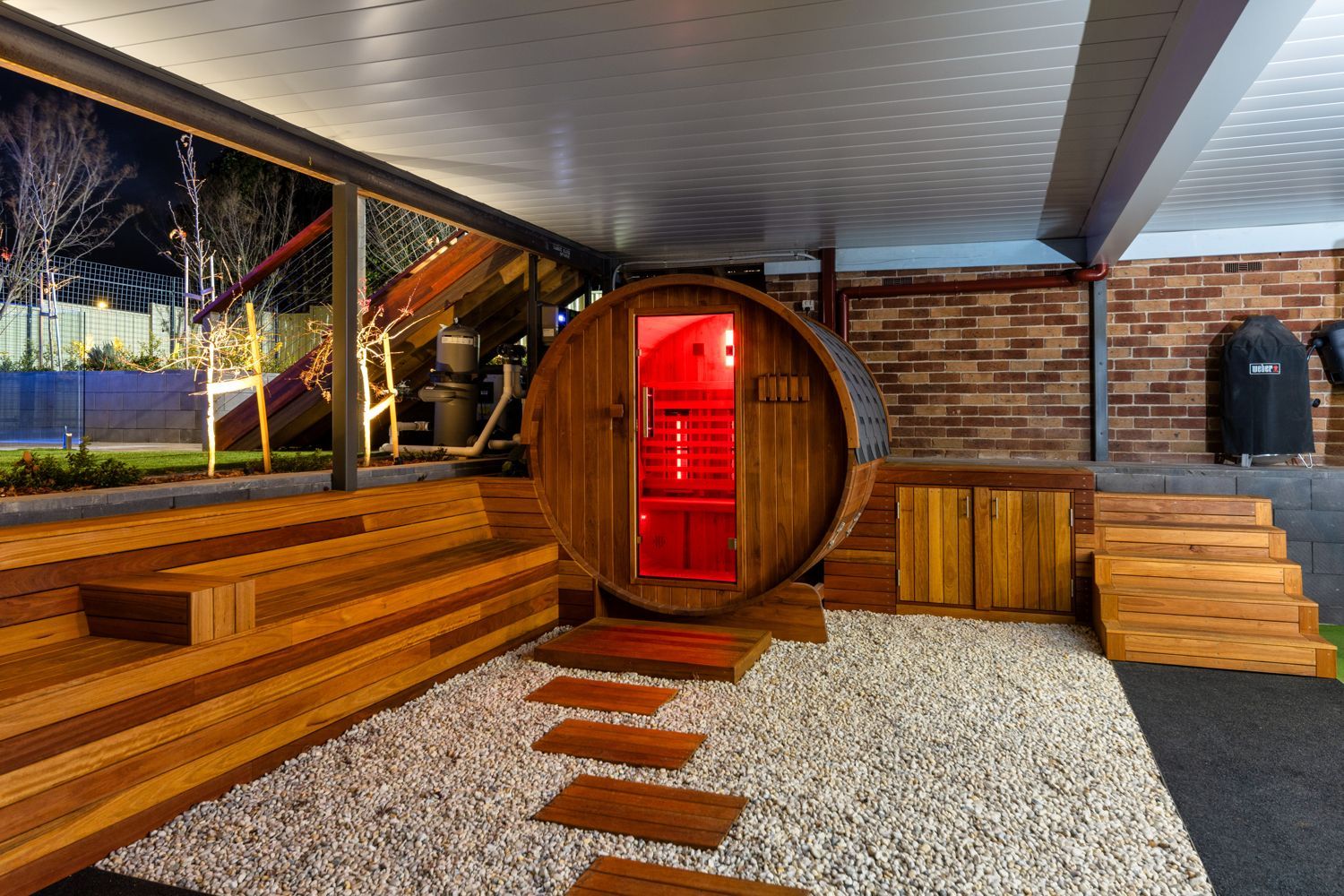 A barrel sauna with a glowing red interior sits on a gravel patio with wood steps under a covered outdoor space.