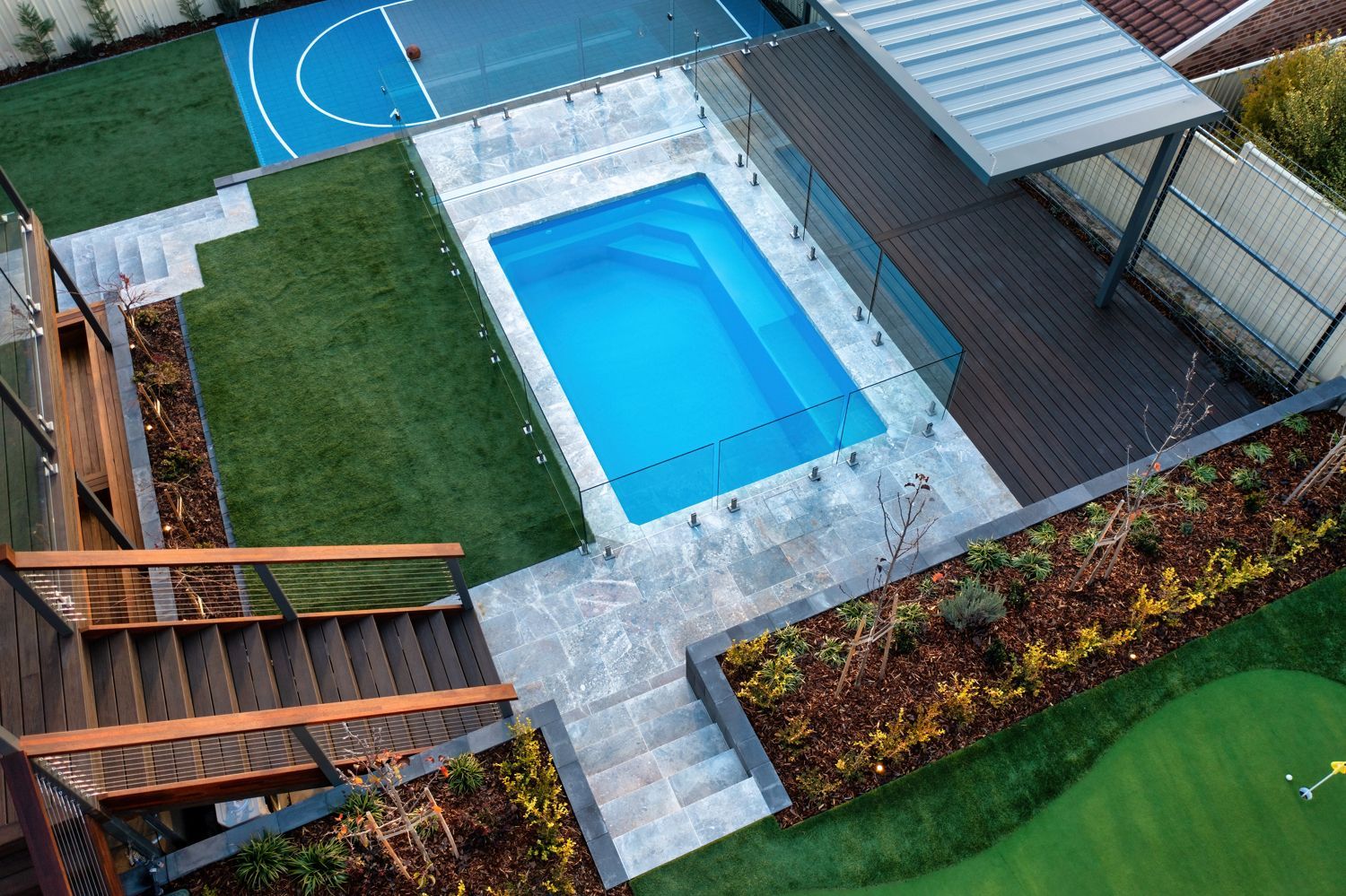 High-angle shot of a backyard with a rectangular swimming pool, turf, a basketball court, and a covered wooden deck.