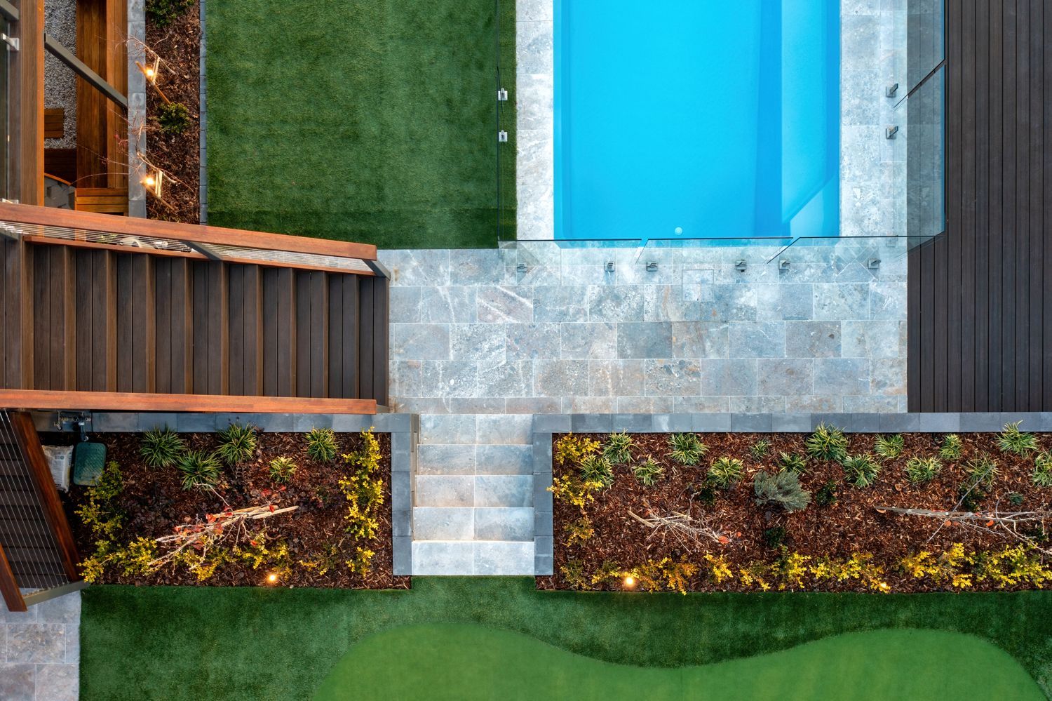 Aerial view of a backyard with a blue rectangular pool, stone patio, wooden stairs, and landscaped garden beds.