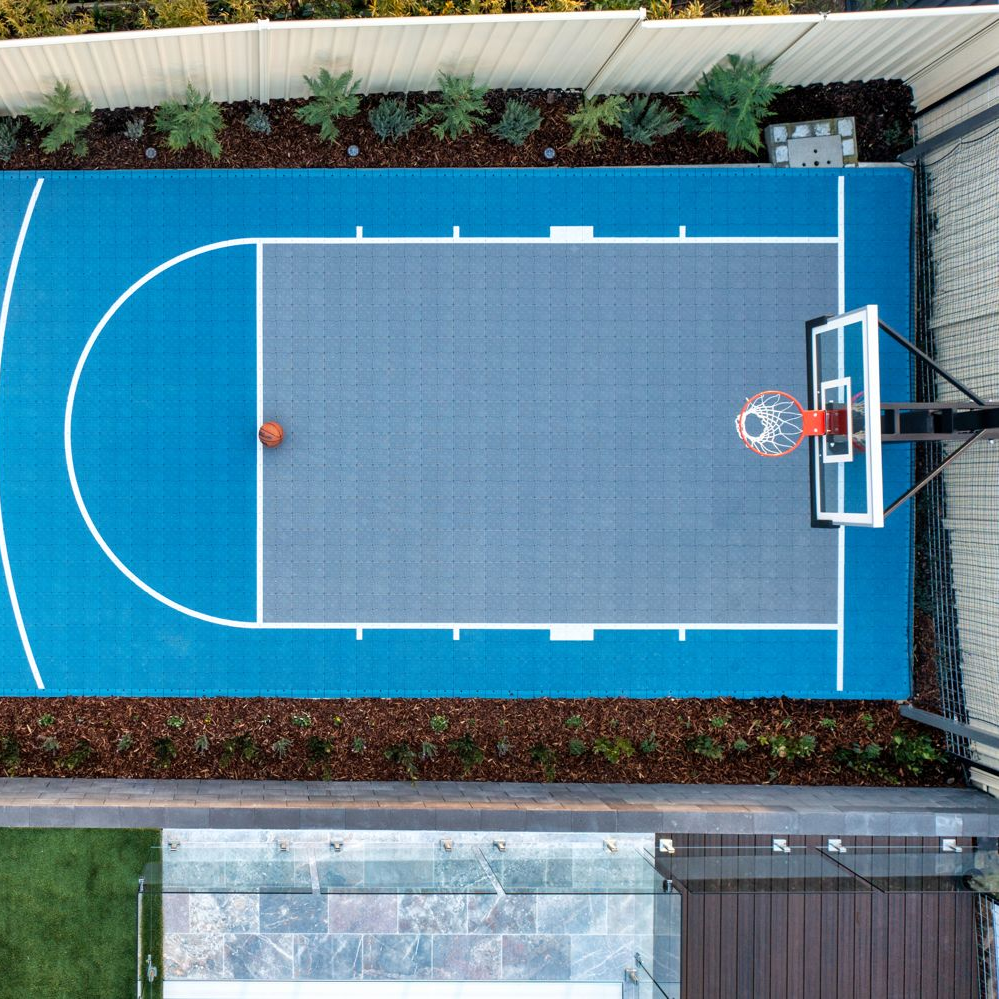 An overhead view of a blue and grey modular backyard basketball court with a hoop and a basketball on the ground.