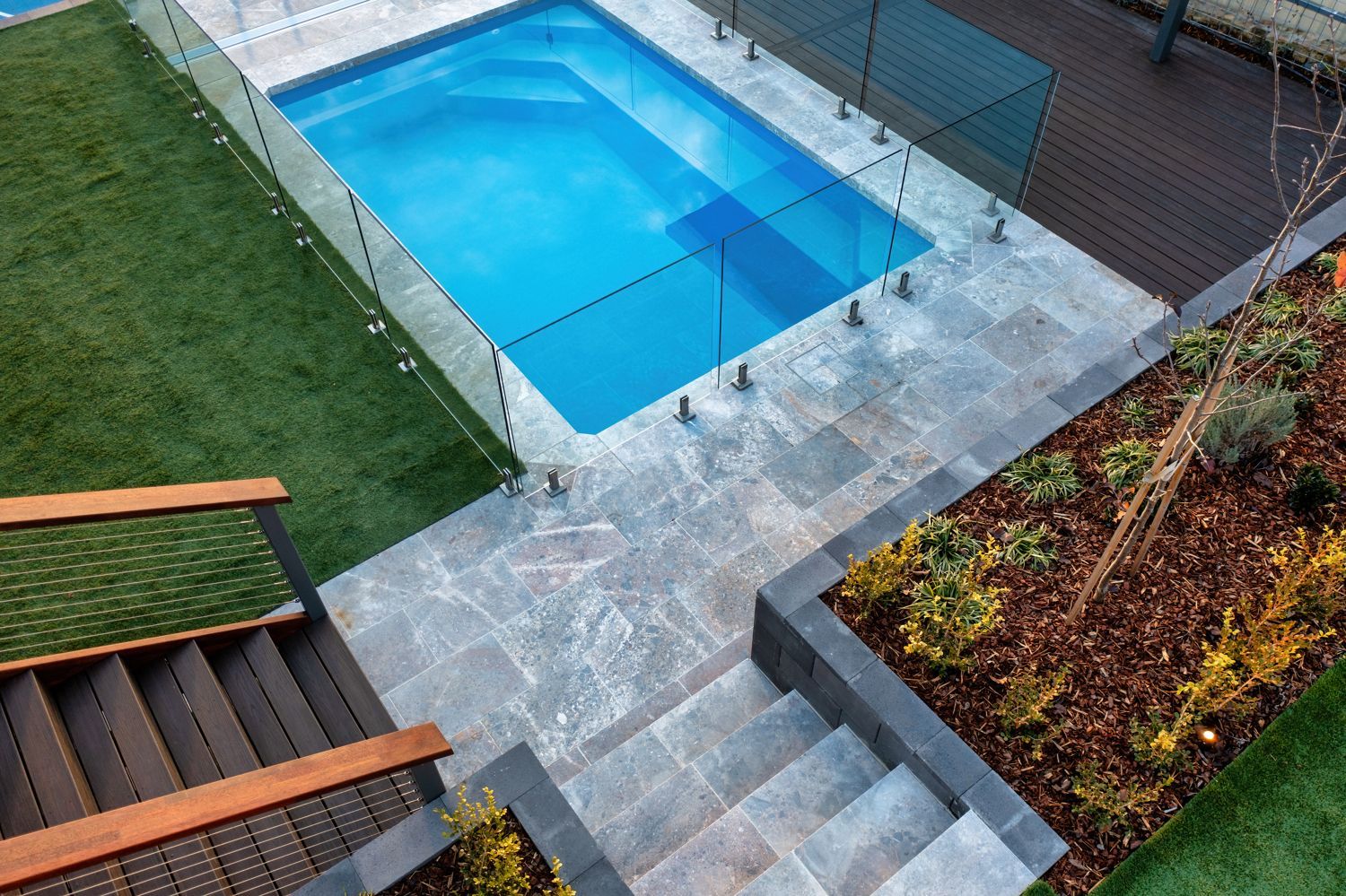 A blue rectangular swimming pool surrounded by grey stone pavers, a wooden deck, stairs, and a landscaped garden bed.