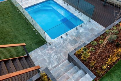 A blue rectangular swimming pool surrounded by grey stone pavers, a wooden deck, stairs, and a landscaped garden bed.