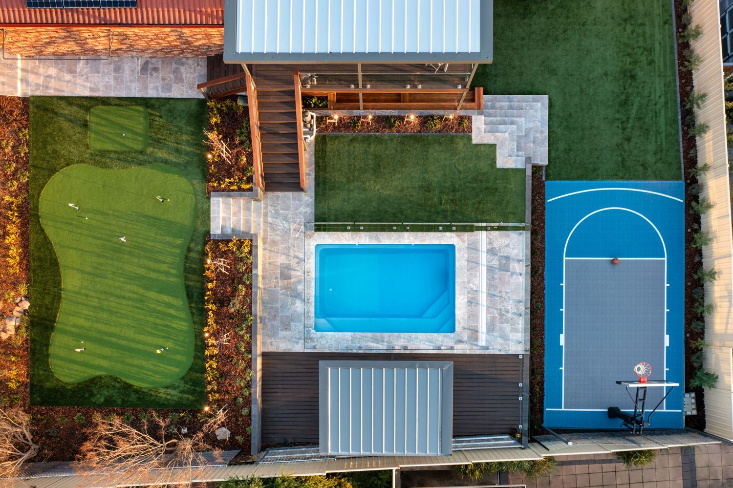 Aerial view of a backyard featuring a putting green, swimming pool, basketball court, and modern house structure.